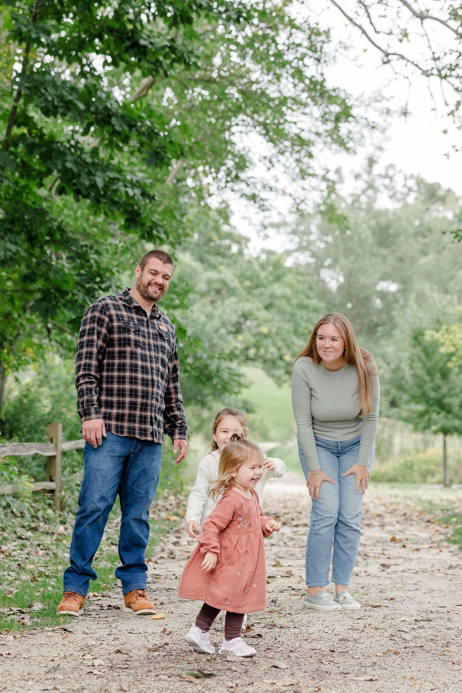 Family of four smiling on a tree-lined gravel path in autumn, with two young girls in front.