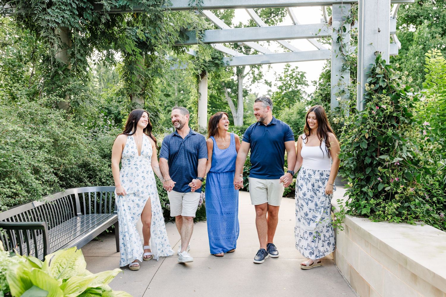 Family of five walks hand-in-hand along lush green garden pergola pathway together.