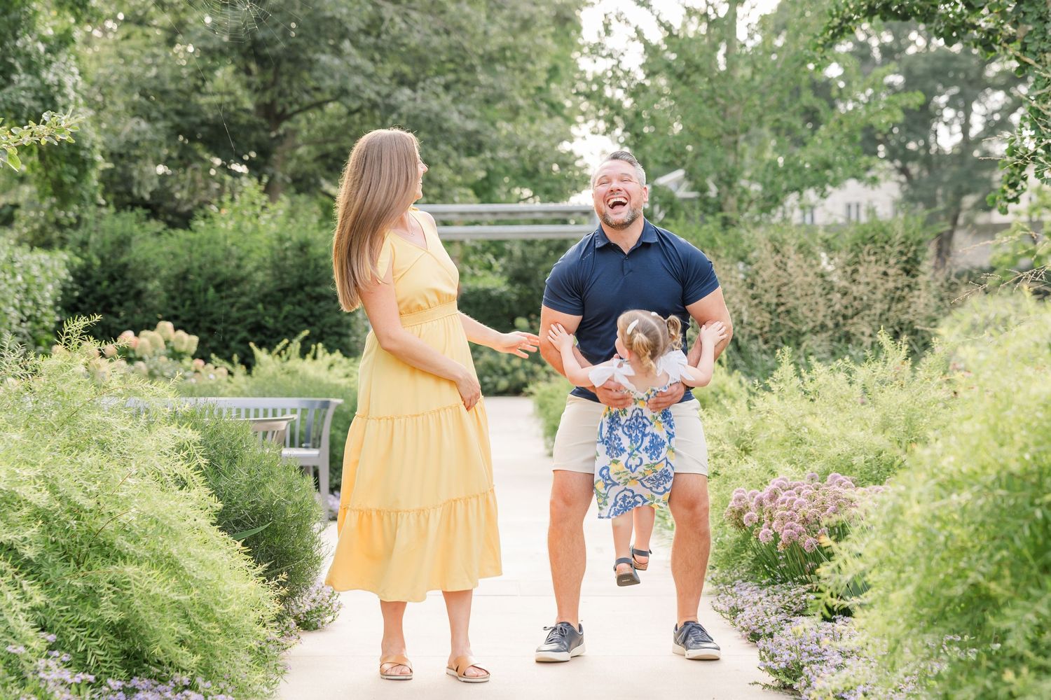 Happy family with toddler girl swinging outdoors in a lush garden path.