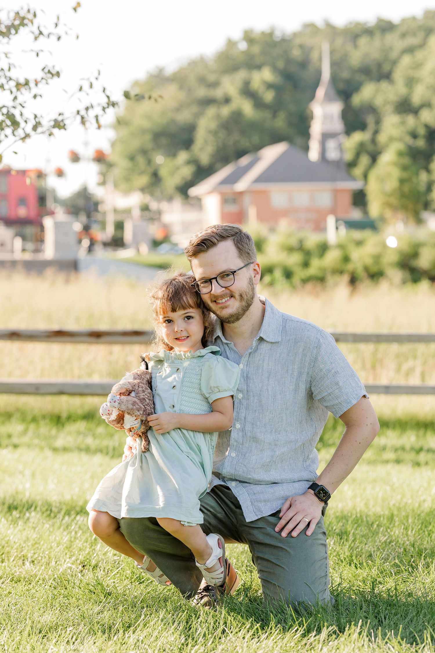 A smiling father kneels on green grass beside his young daughter in a mint dress during an outdoor photo session.