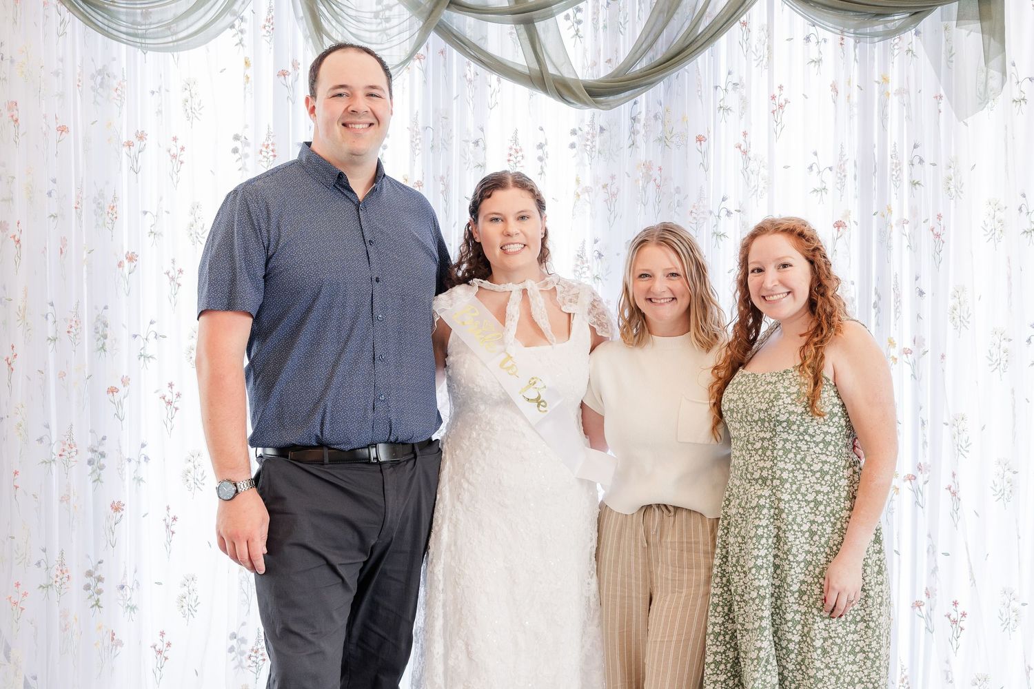 Four people posing together smiling in front of a sheer floral curtain at a bridal shower event.