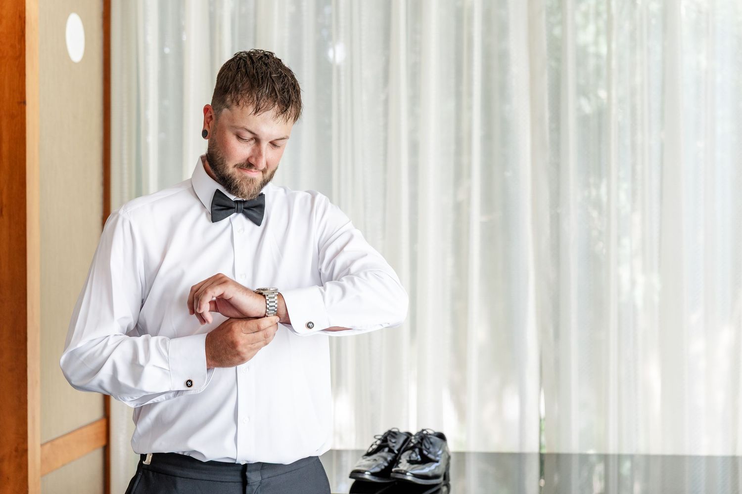 Groom in white shirt and bow tie checking watch while getting ready on wedding day.