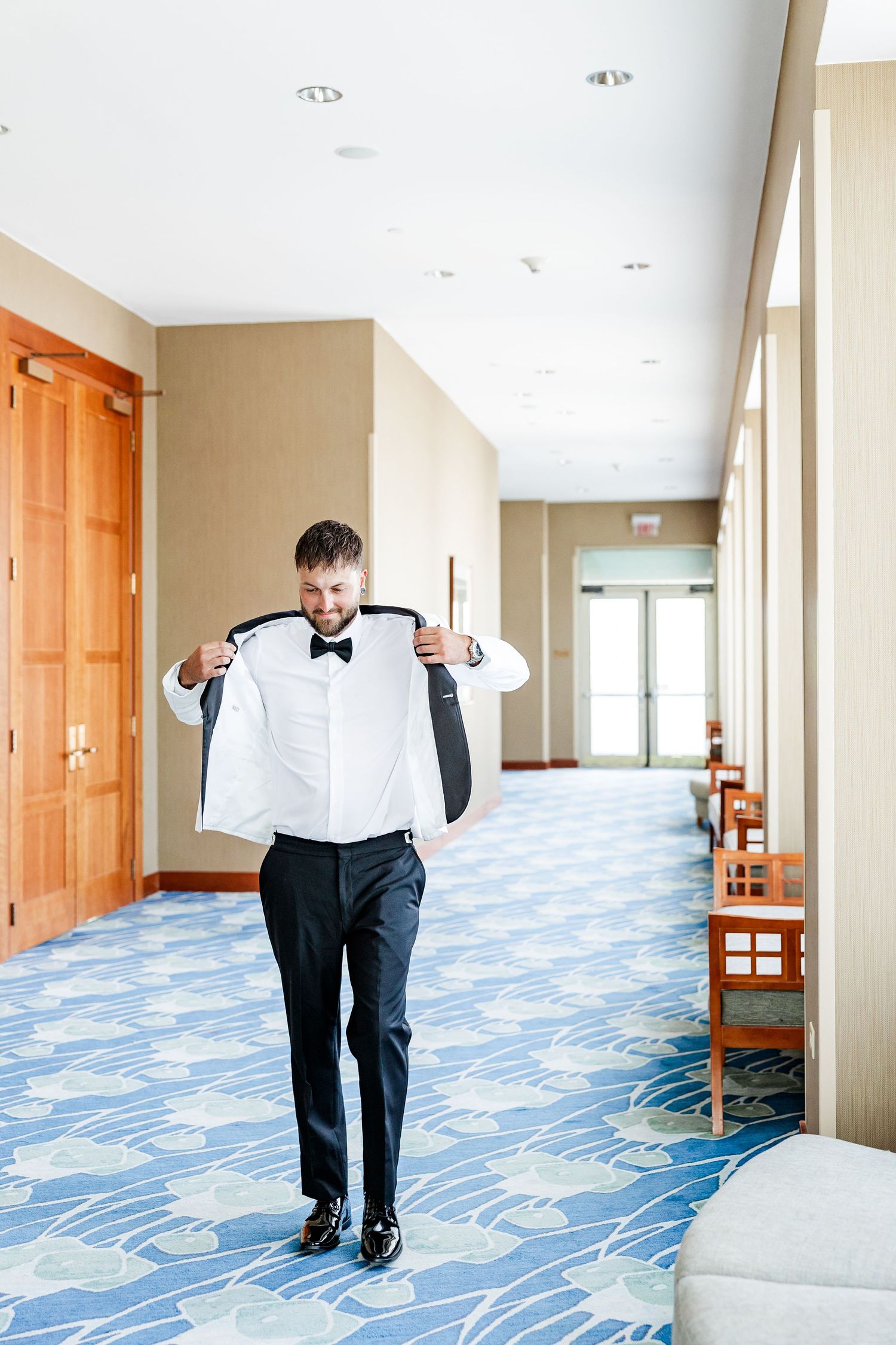 Groom in white tuxedo putting on jacket in elegant hotel corridor with blue patterned carpet.