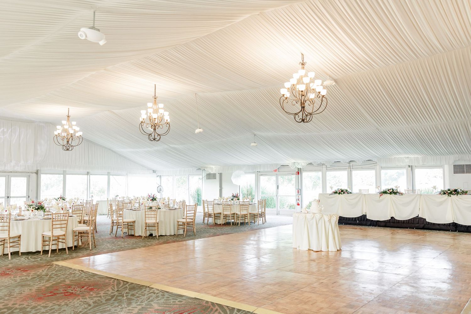 Elegant wedding reception tent with draped white ceiling, crystal chandeliers, round tables, and a spacious dance floor.