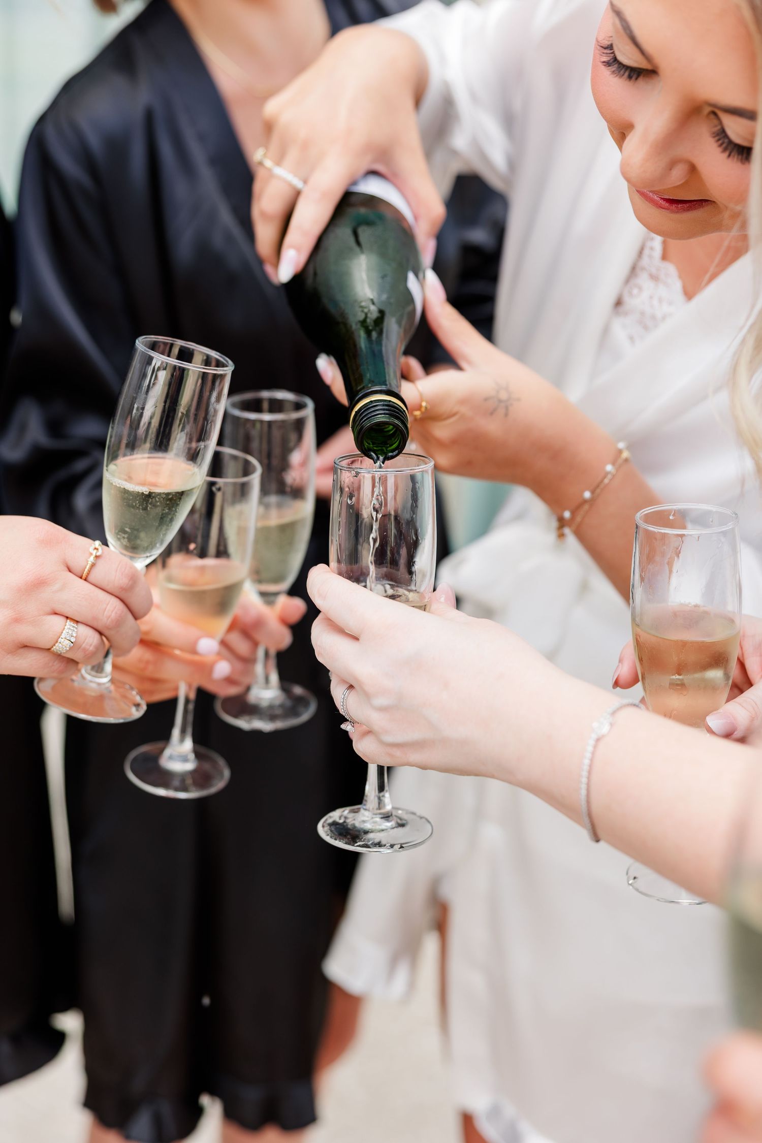 Bride pouring champagne into glasses held by bridesmaids during wedding morning preparations.