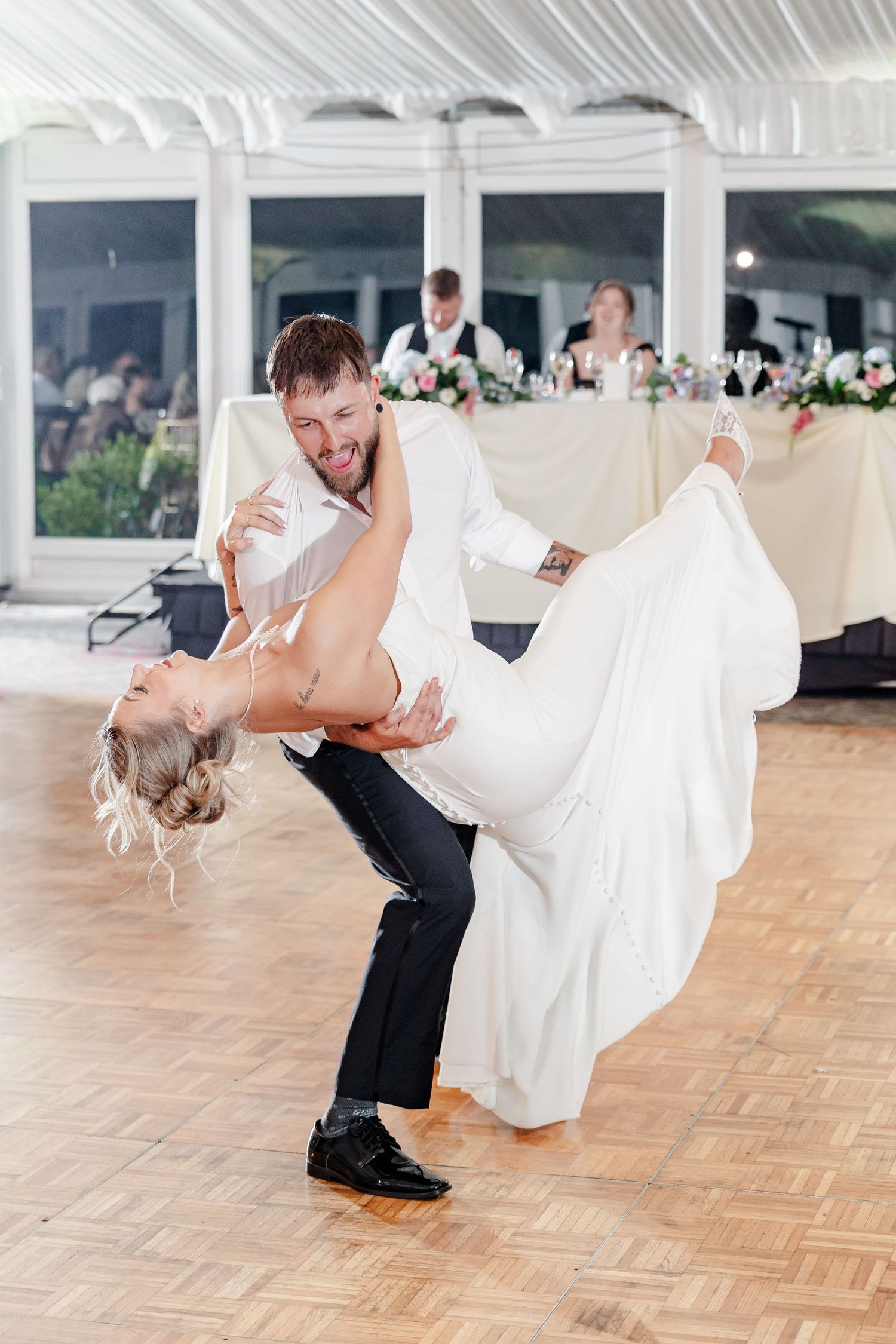 Groom dips bride during first dance at elegant wedding reception with guests seated in background.