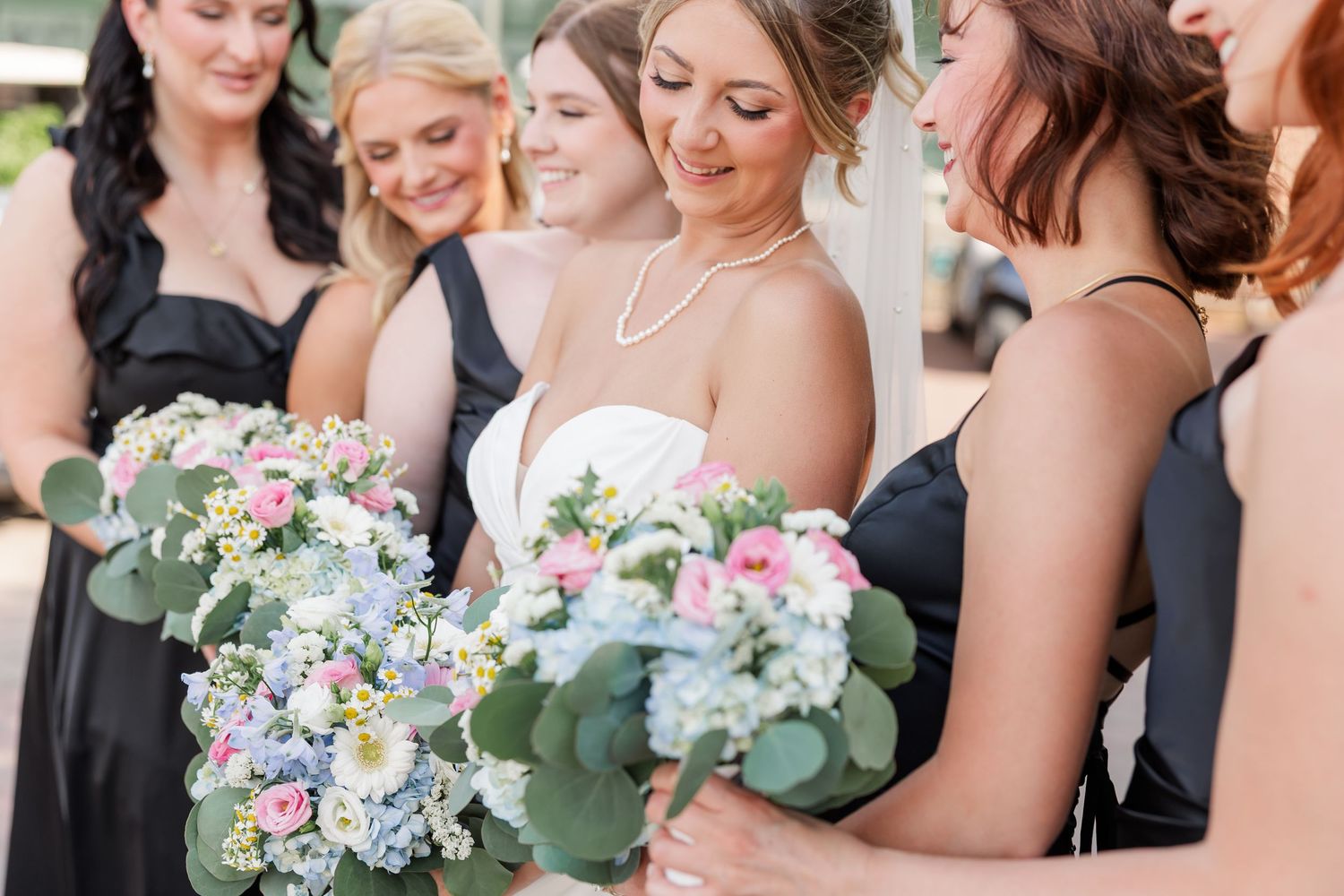 Bride in white gown with pearl necklace surrounded by bridesmaids in black holding floral bouquets.