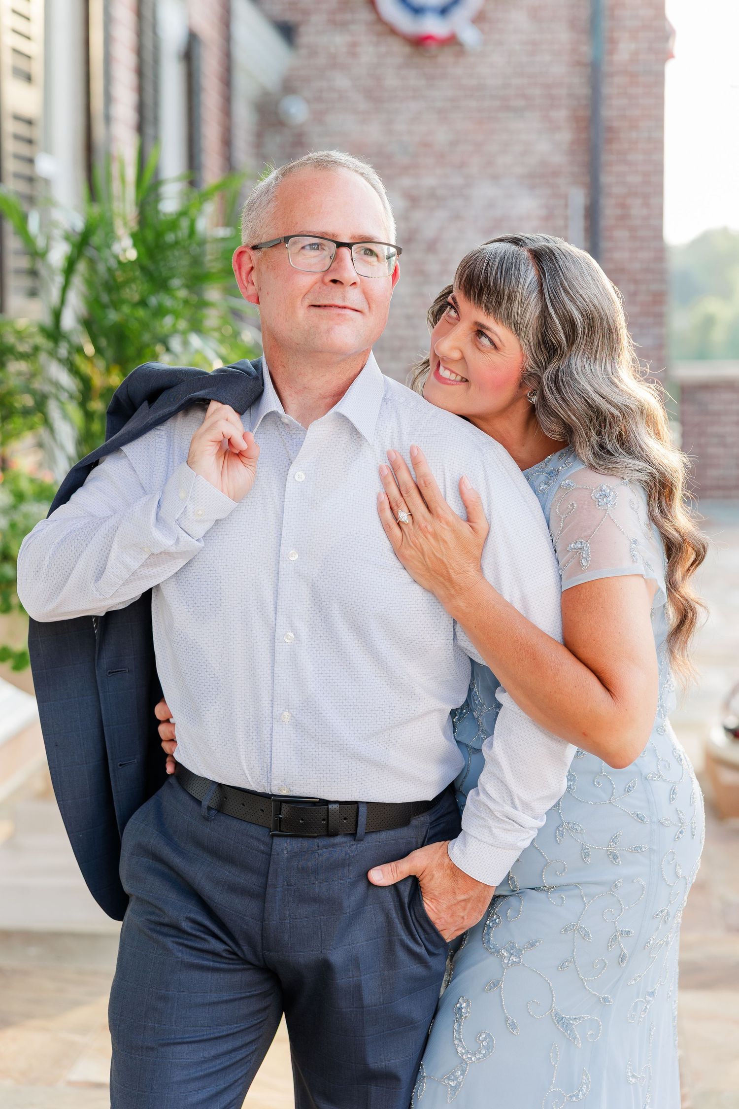 Couple posing outdoors, man in white shirt holding jacket over shoulder, woman in blue dress embracing him.