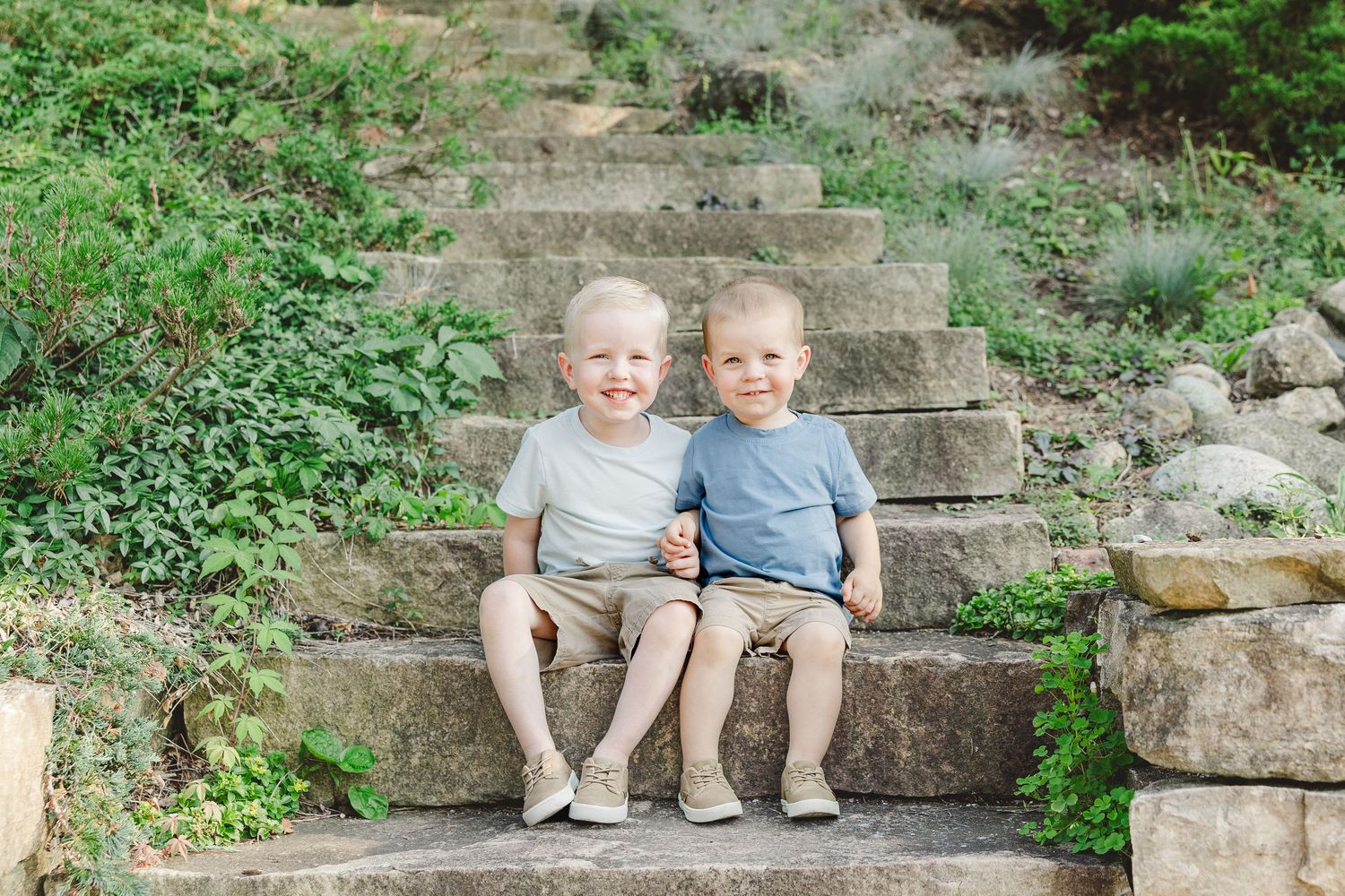 Two young children in matching blue shirts sit together on stone garden steps surrounded by greenery.