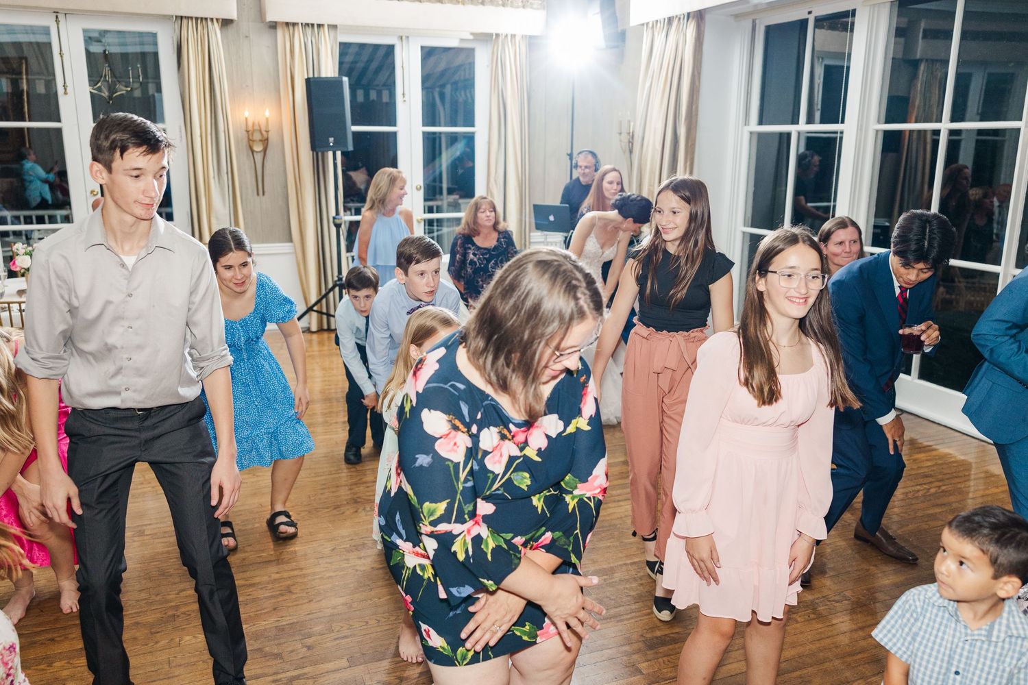 Party guests dance and celebrate in an elegant room with large windows and curtains.