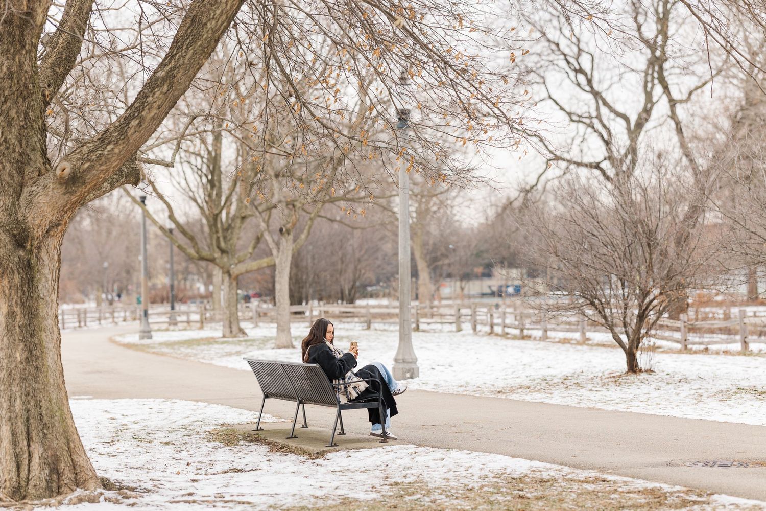 A person sits on a park bench surrounded by snow-covered ground and bare winter trees in a serene setting.