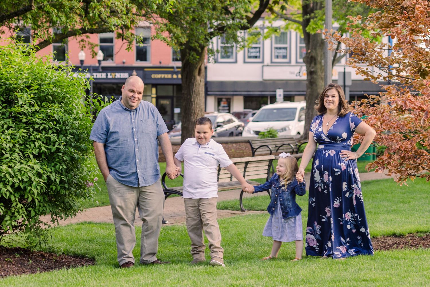 Family stands together holding hands on grass with historic buildings and trees in background.