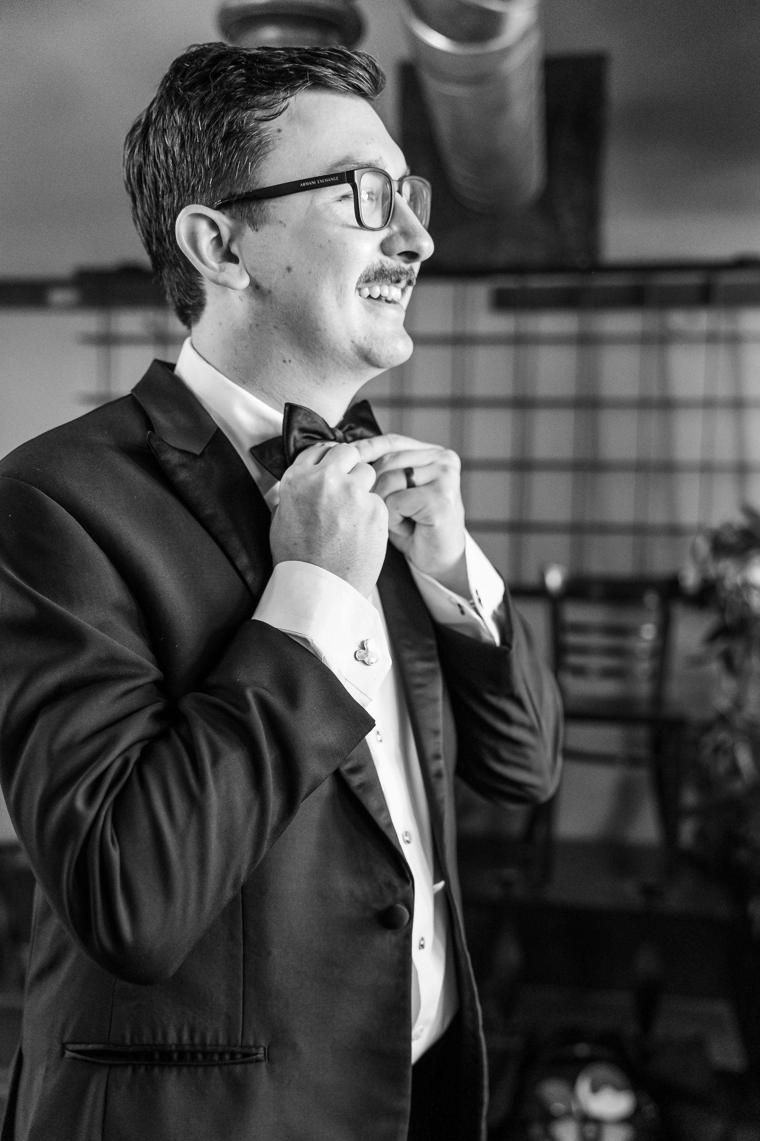 Side profile of person in tuxedo and glasses adjusting bow tie while smiling.