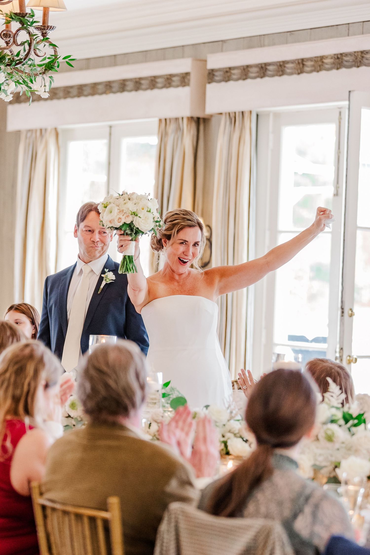 Joyful celebration scene at an elegant indoor wedding reception with white decor and natural lighting.