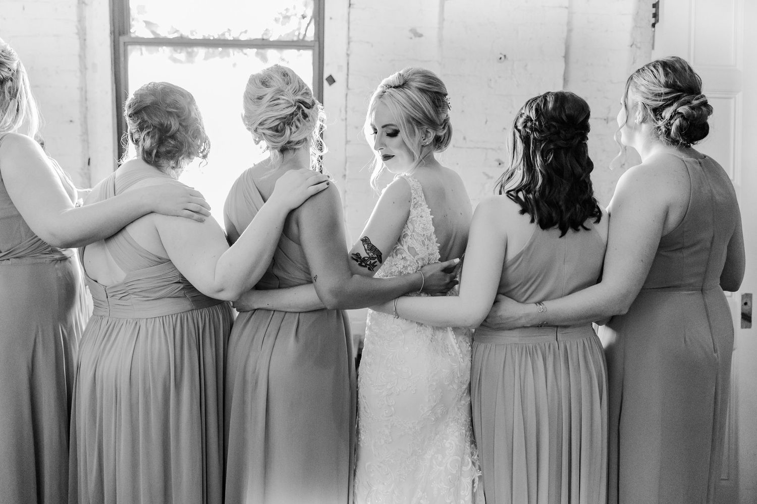 Black and white image of bridal party embracing while facing window in rustic setting.