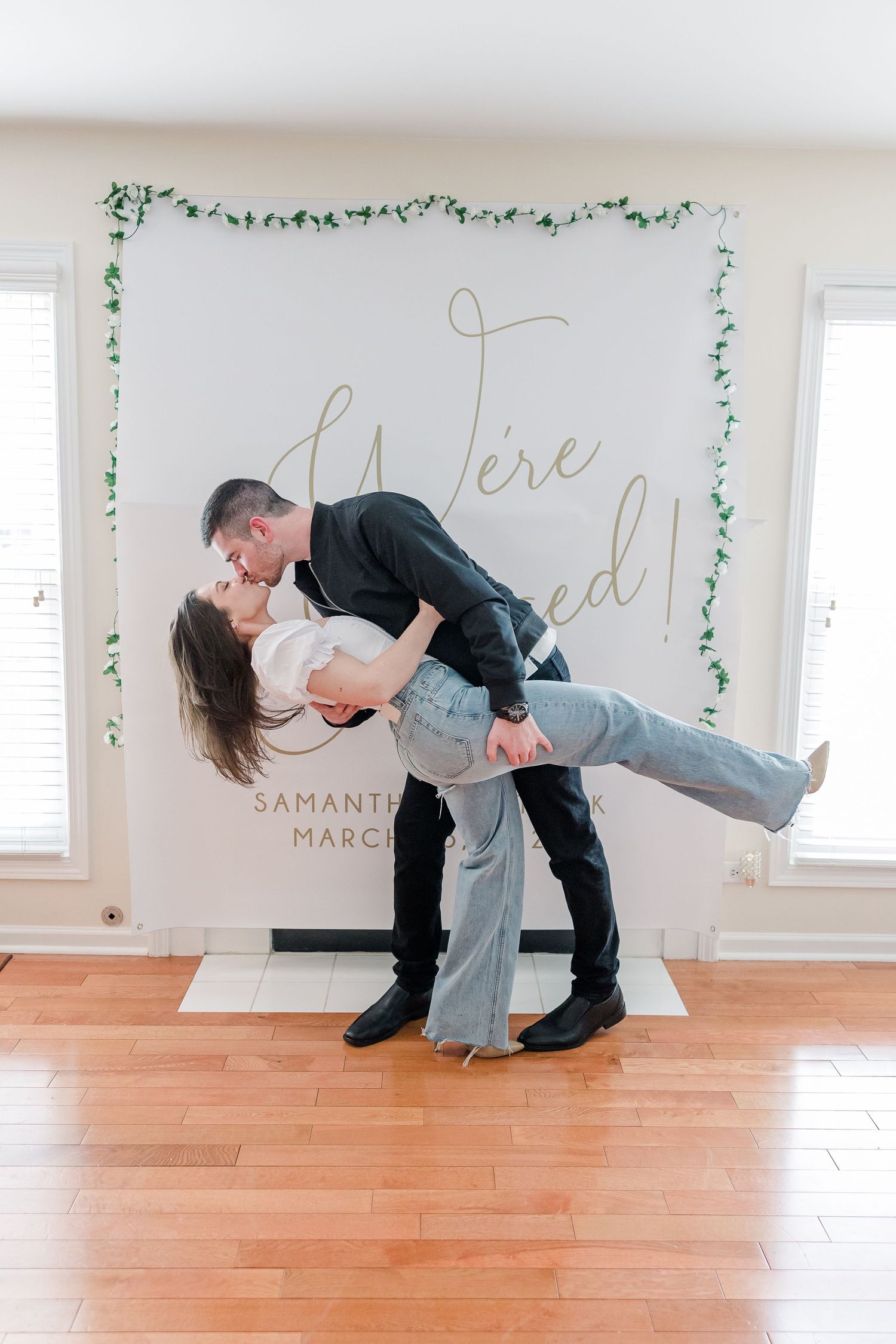 Couple in casual attire sharing romantic dip pose against decorated engagement backdrop.