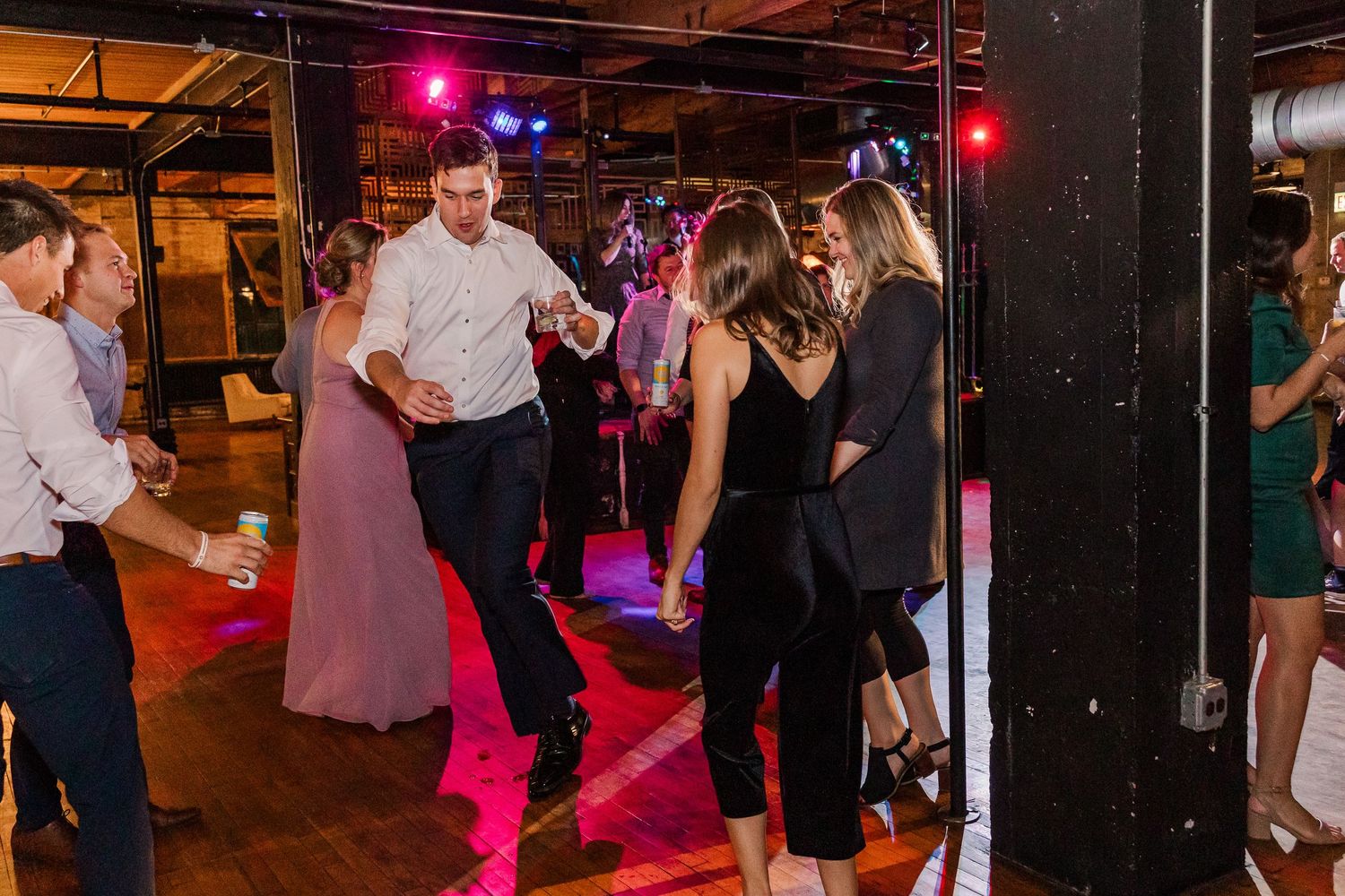 People dancing under pink lighting at a wedding reception in an industrial venue.