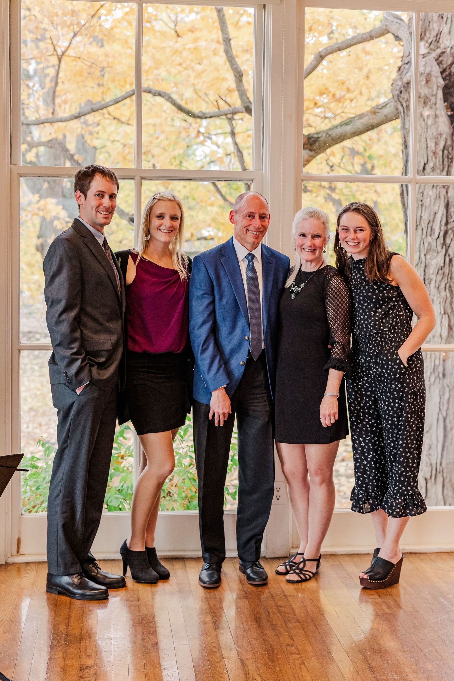 Formal group photo of five people standing together by a window with autumn foliage visible outside.