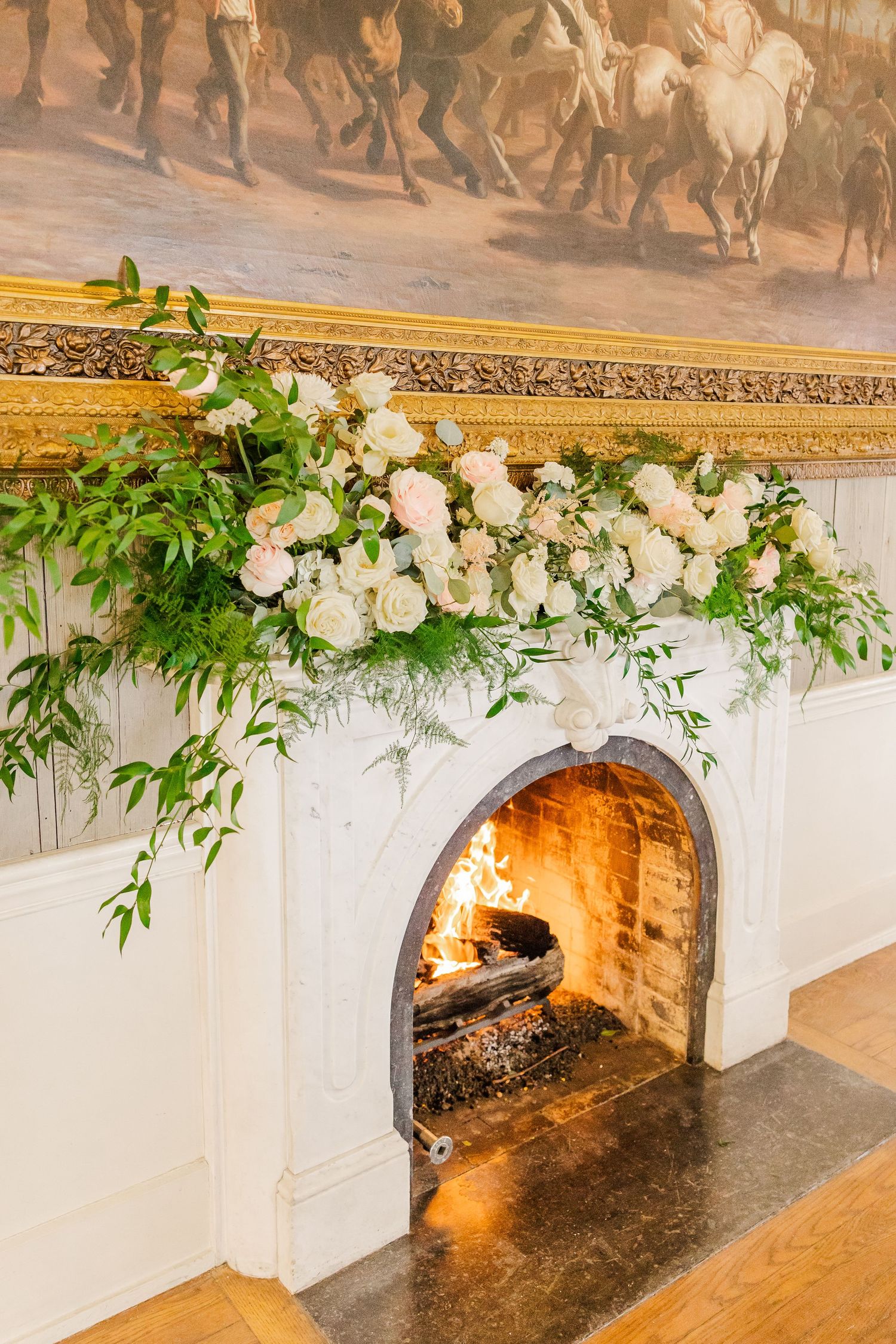 White floral arrangement adorning fireplace mantel with lit fire below and equestrian artwork above.