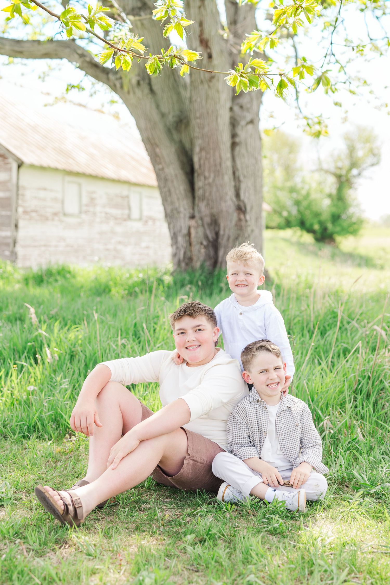 Three boys in casual clothing relax under large tree near rustic barn.