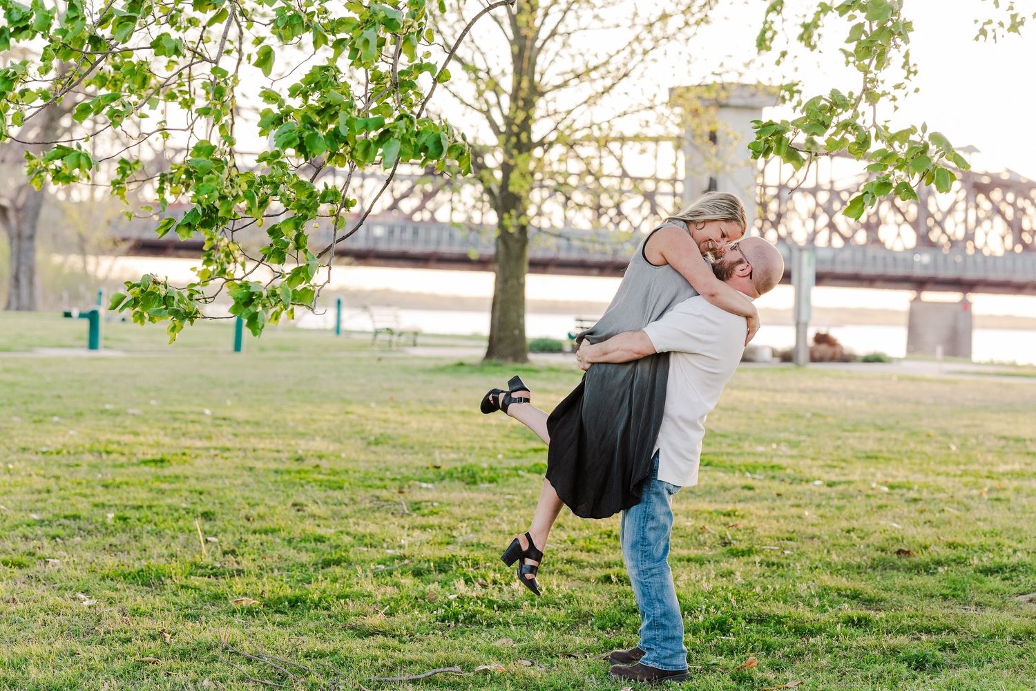 A playful couple shares a romantic lift pose in a park with a bridge visible behind them.