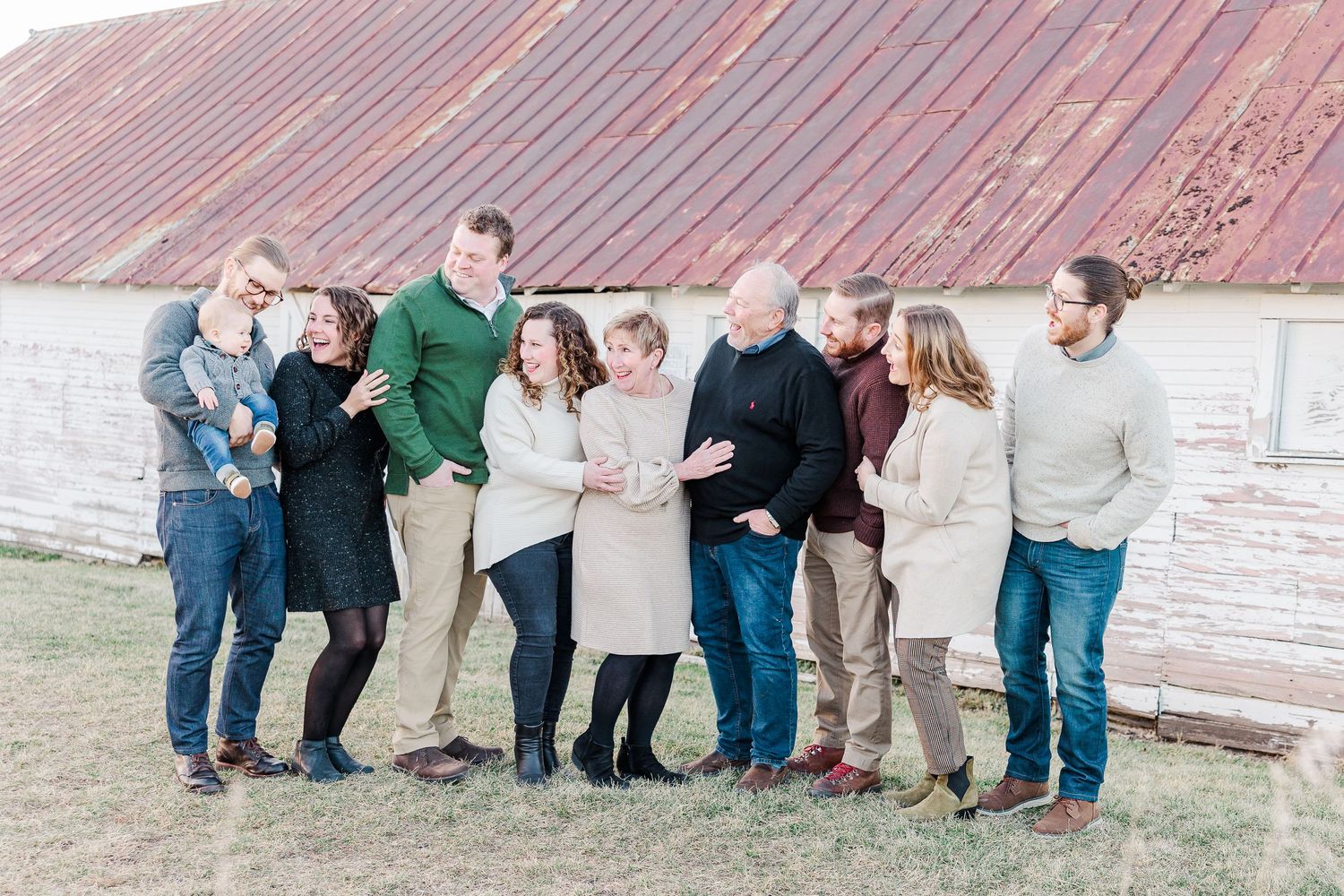 A large family group poses together in casual clothing against a rustic barn backdrop.