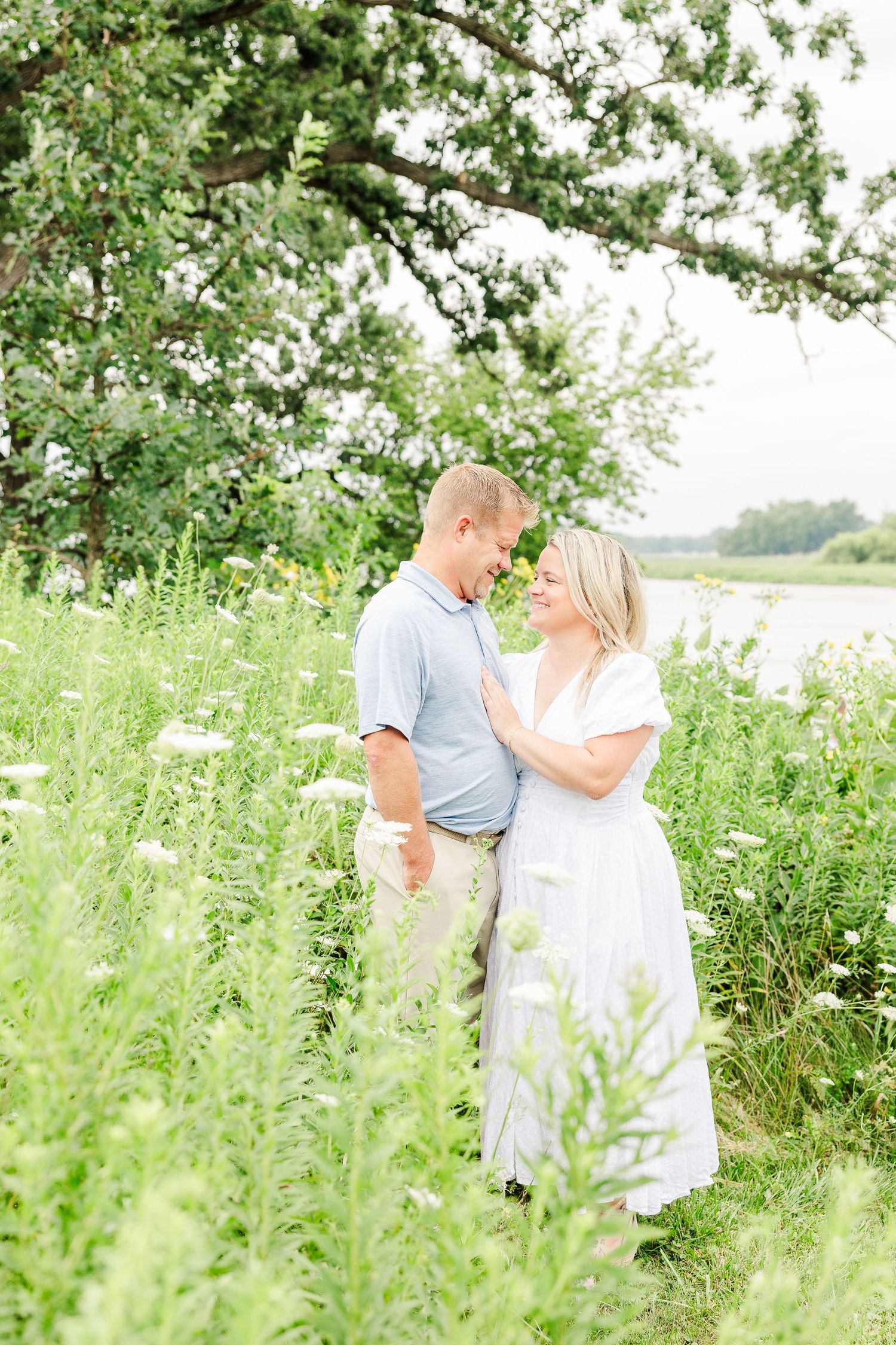 A romantic couple standing among wildflowers in a sunny meadow by the water.