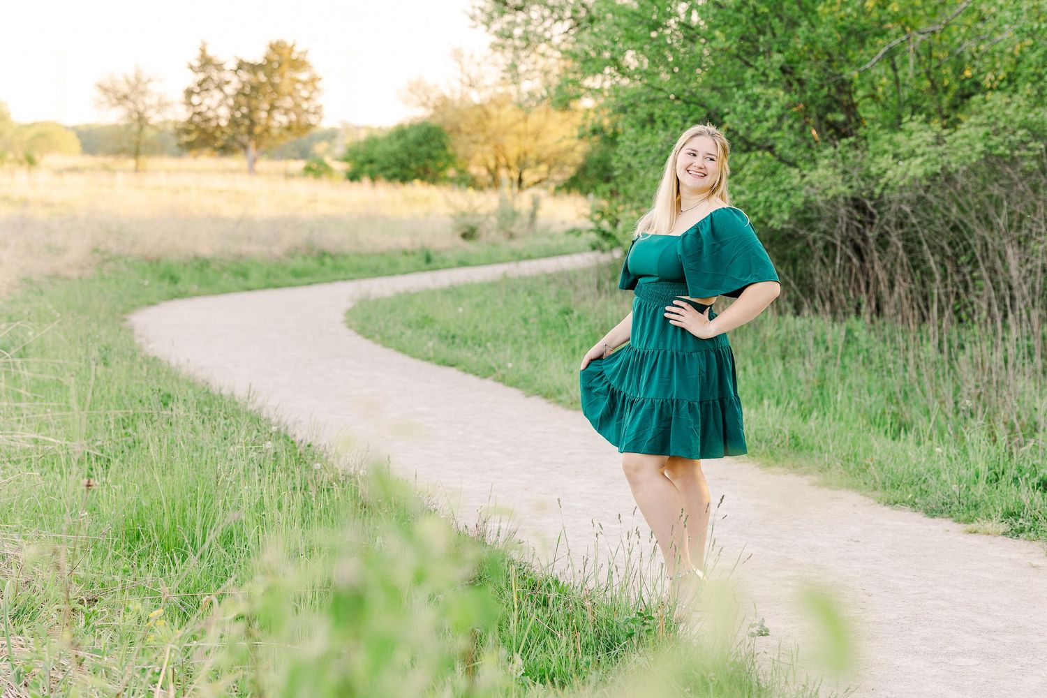 A person in an emerald green dress poses on a winding path through spring meadows.