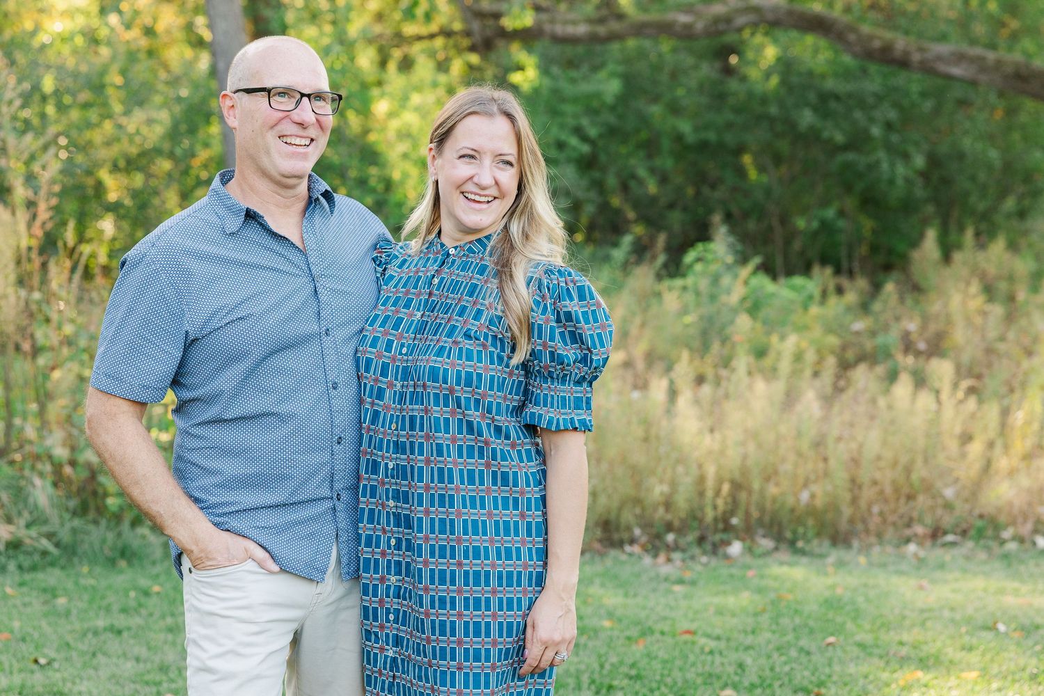 A couple stands together outdoors in coordinating blue outfits against a natural green background.