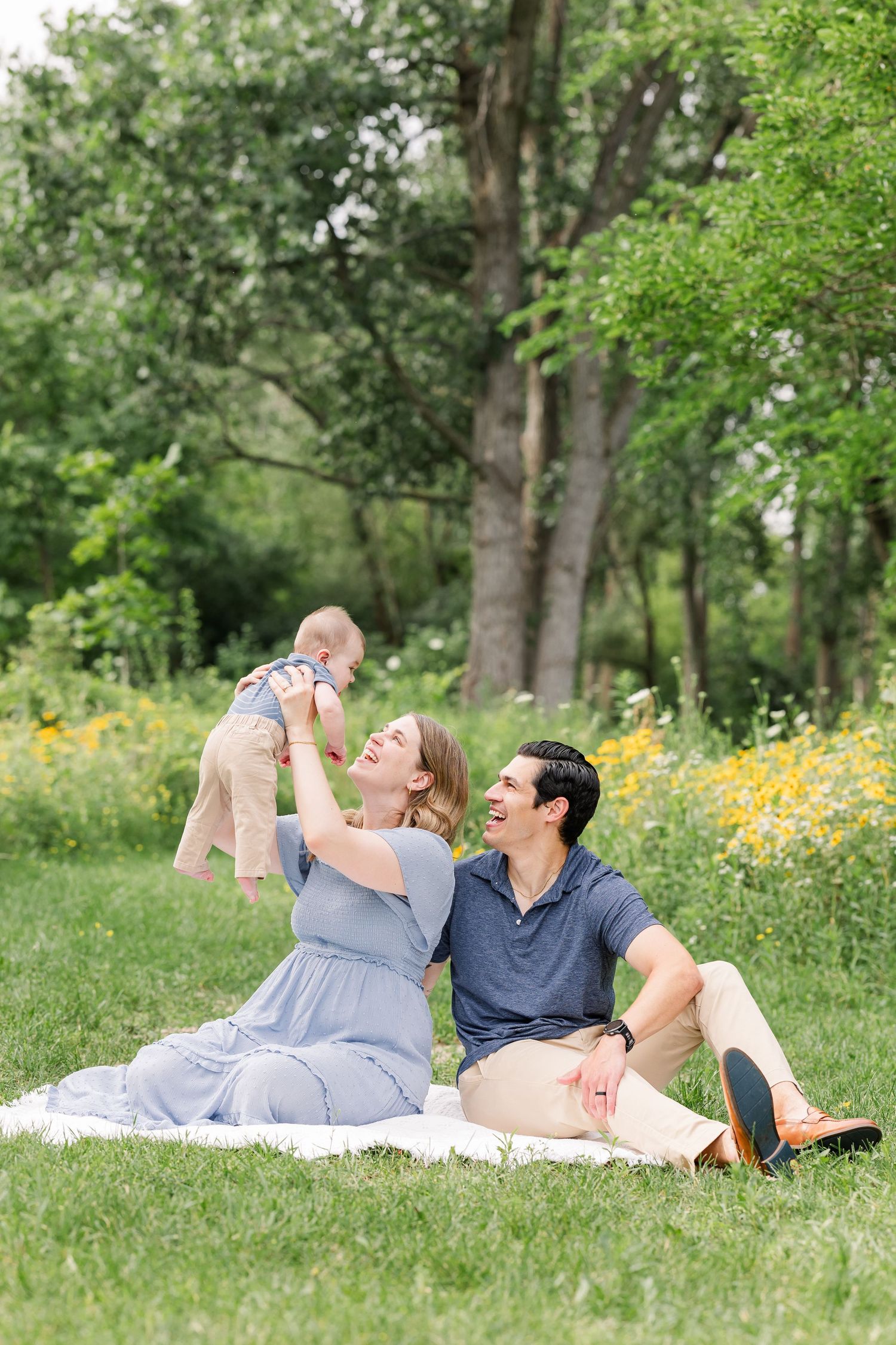 Happy couple sitting on blanket in park, mom lifting baby while dad laughs beside her.