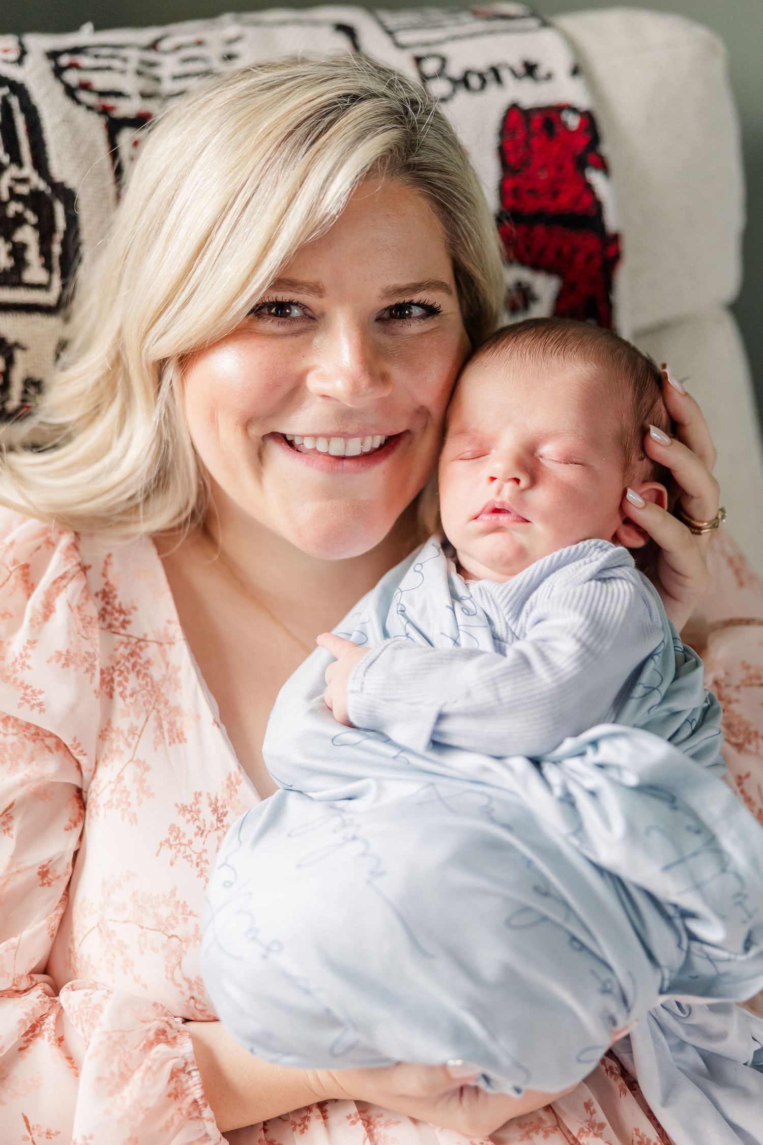 Happy blonde mother in pink dress cuddling sleeping newborn baby dressed in light blue against her cheek.