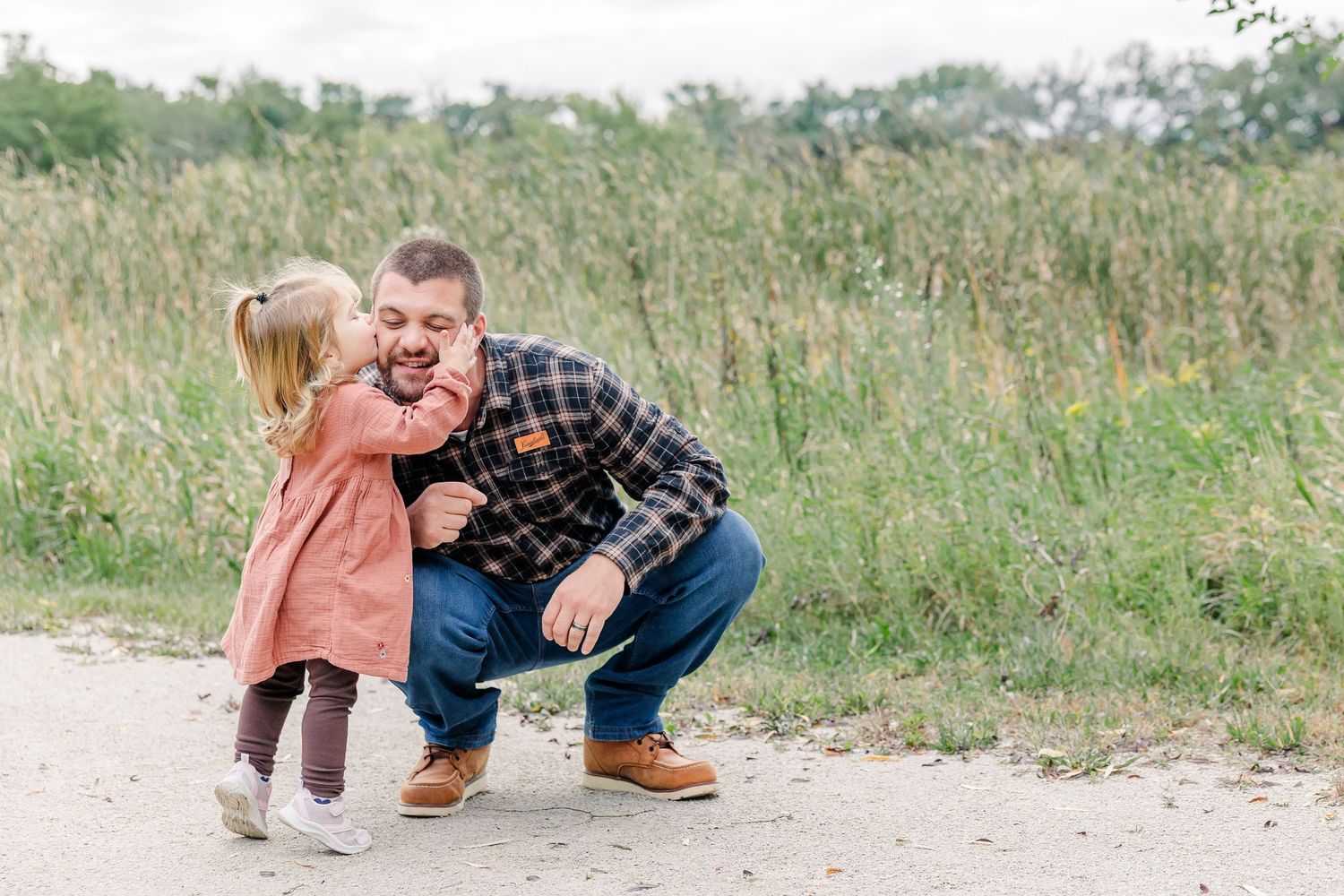 A little girl in a pink dress gives her laughing father a kiss on the cheek outdoors.