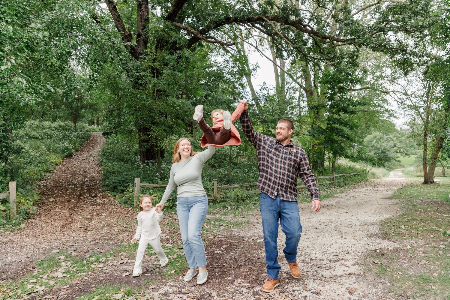 A joyful family of four plays outdoors on a wooded path, with parents swinging their child in the air.