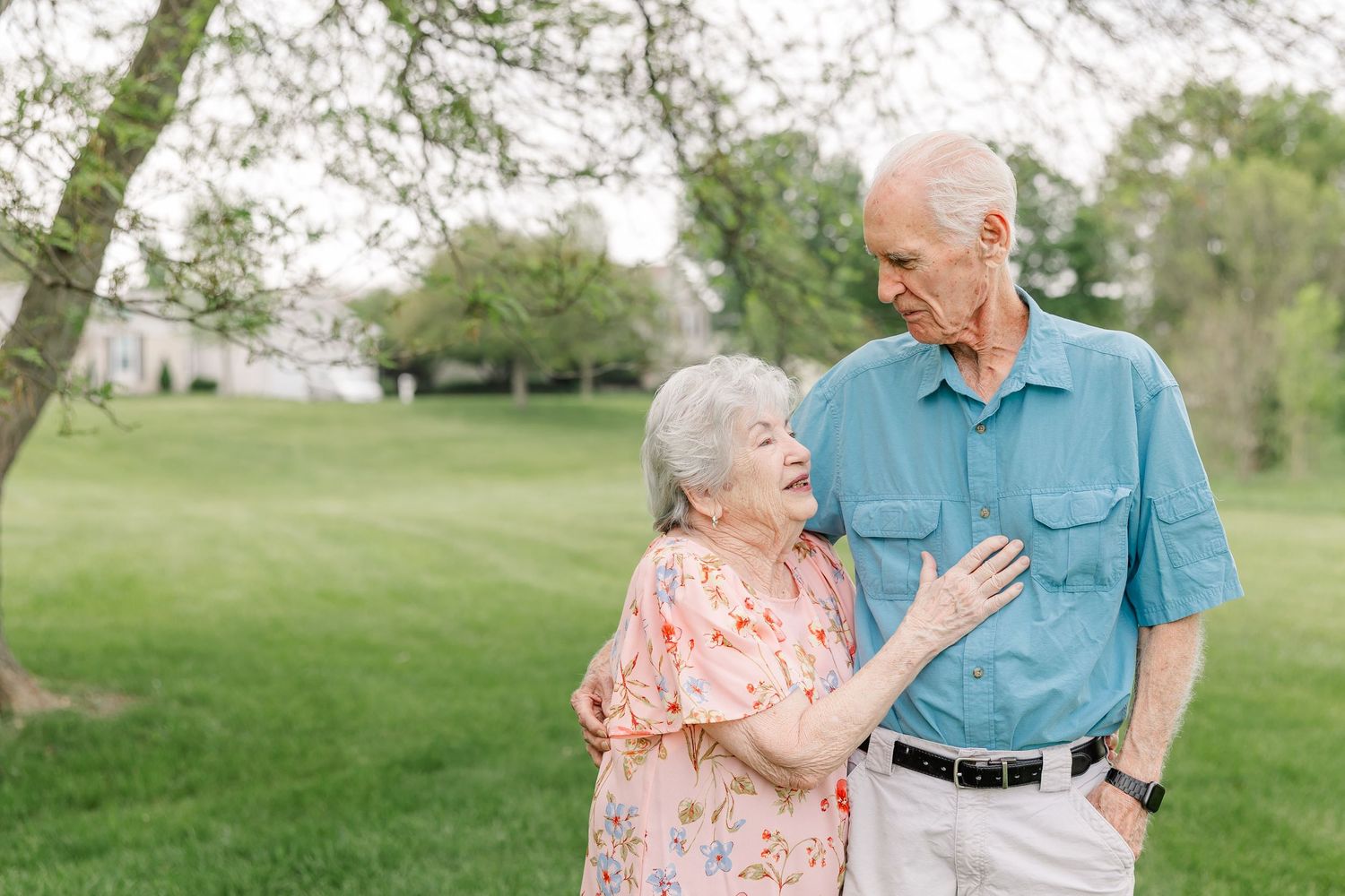 Elderly couple smiles warmly at each other while standing together outdoors beneath a blooming spring tree.