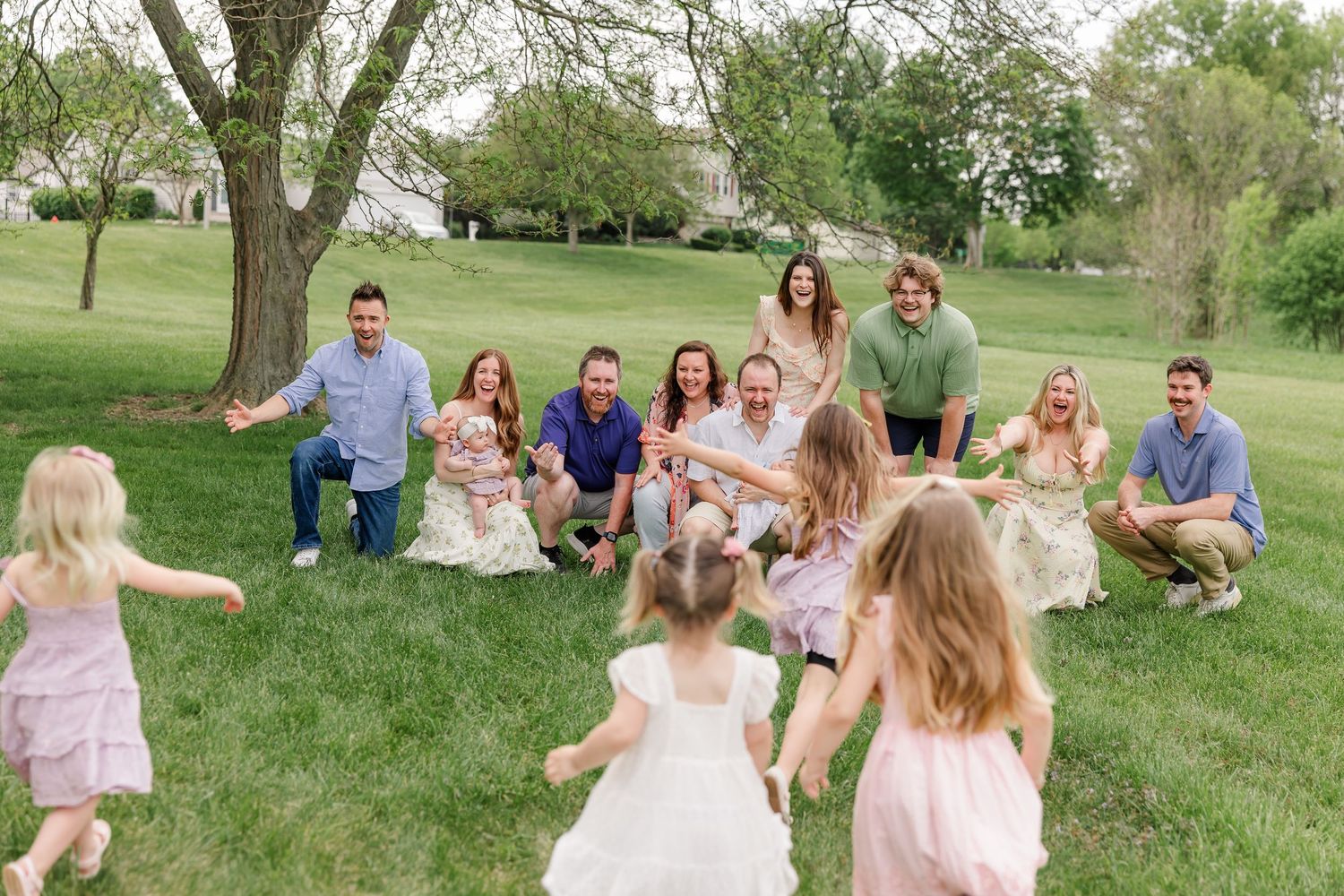 Large extended family gathered outdoors on green grass, laughing as young children run toward them near a tree.