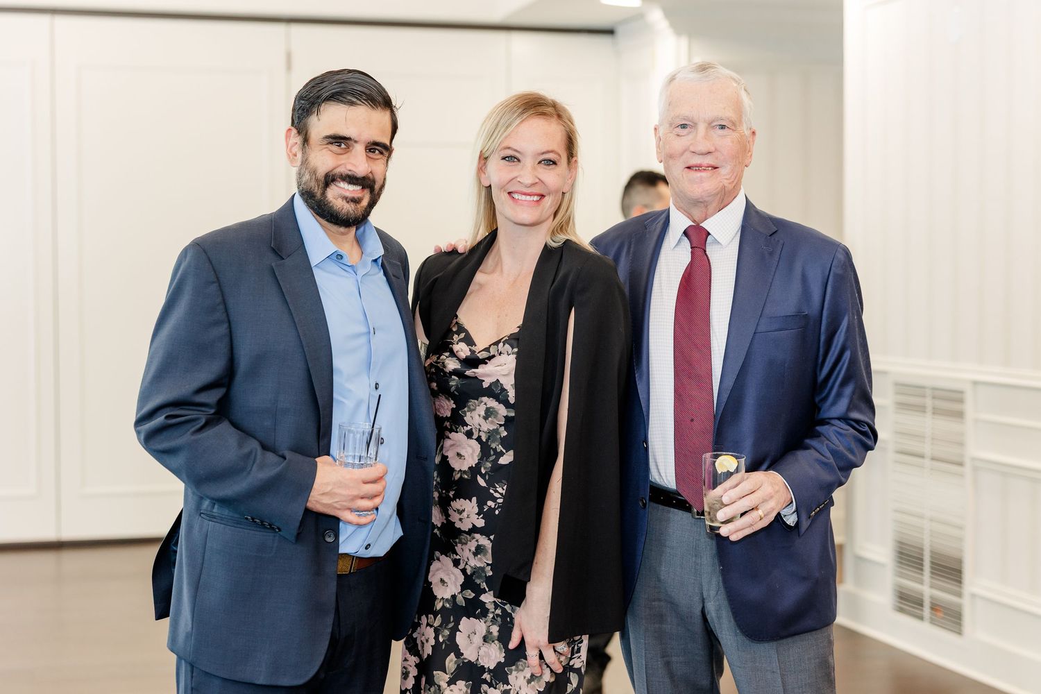 Two men in navy blazers and a smiling woman in a floral dress pose with drinks at a wedding reception.