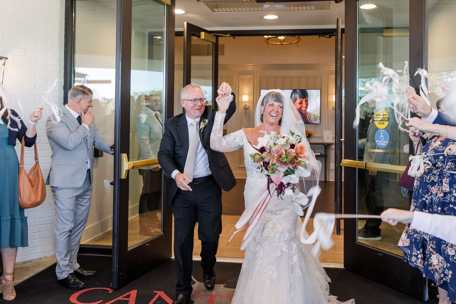 Smiling bride in a white gown holding a bouquet walks hand in hand with a groom through a doorway as wedding guests cheer and wave ribbons.