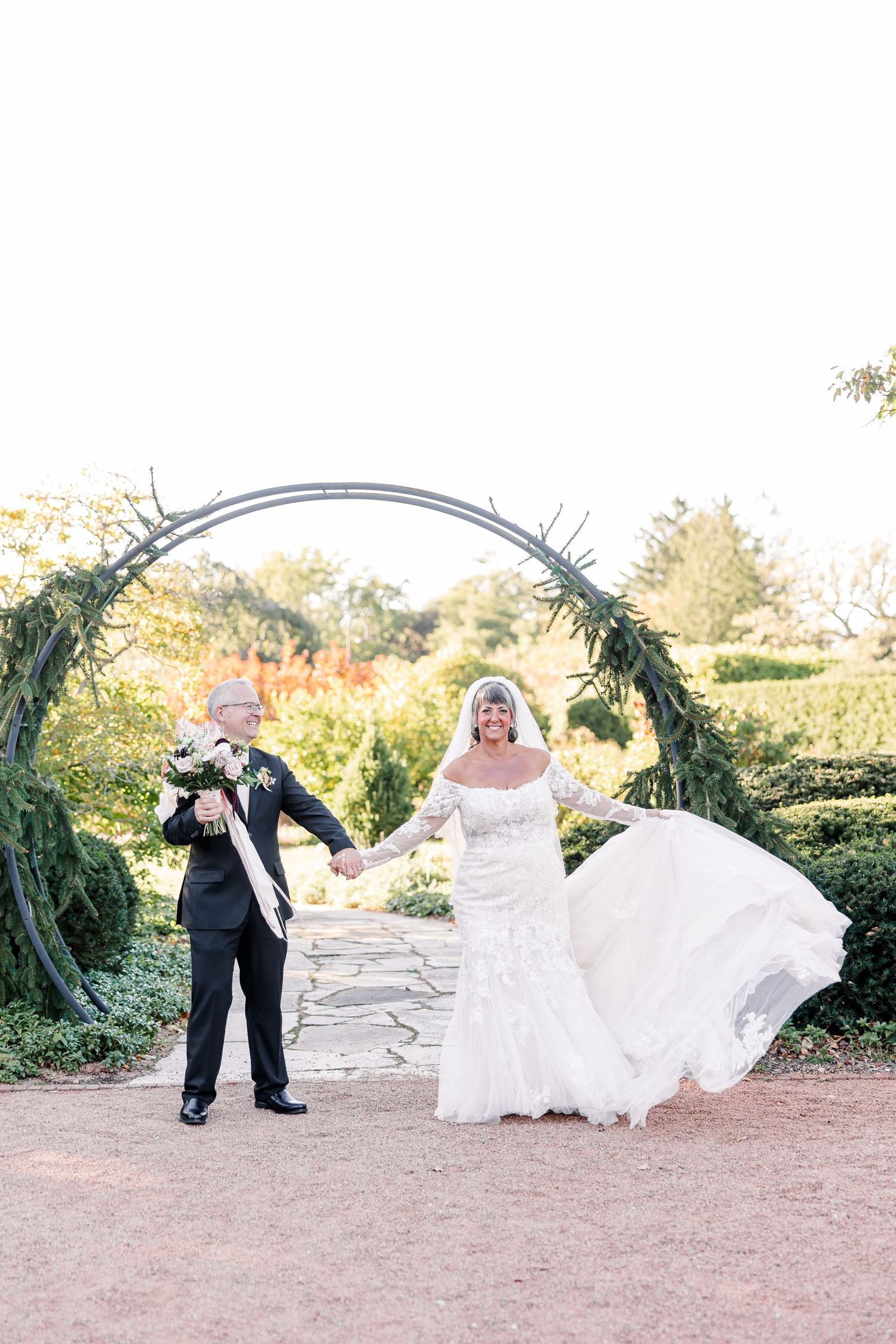 Smiling bride in a lace wedding gown with a flowing skirt holds hands with a groom in a black suit before a greenery arch in a garden.