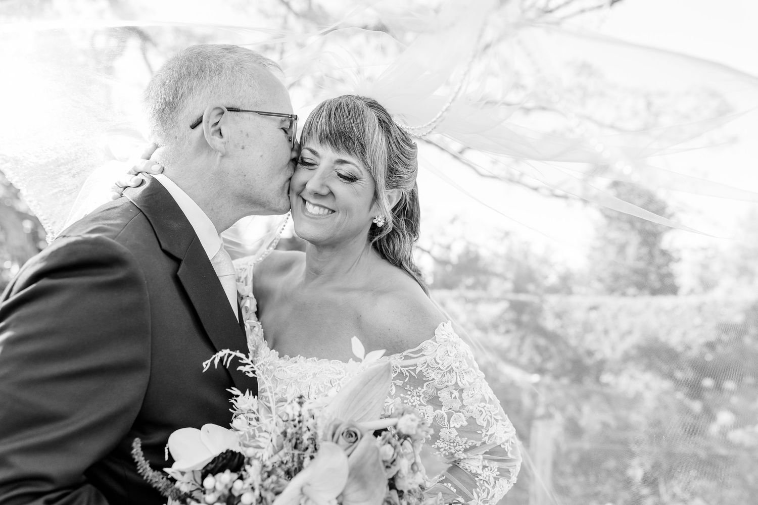Groom in a suit kisses a smiling bride in a lace wedding dress holding a bouquet in a black and white outdoor wedding portrait.