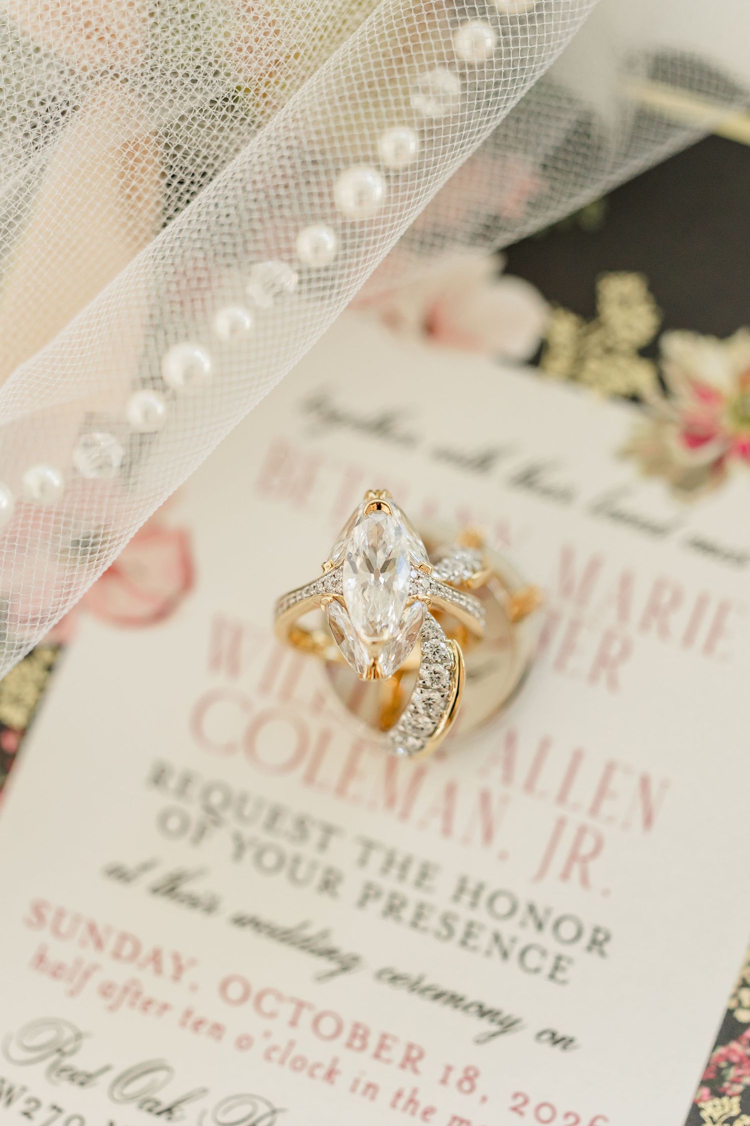 Marquise diamond gold engagement ring with wedding band resting on a floral wedding invitation beneath a pearl-trimmed veil.