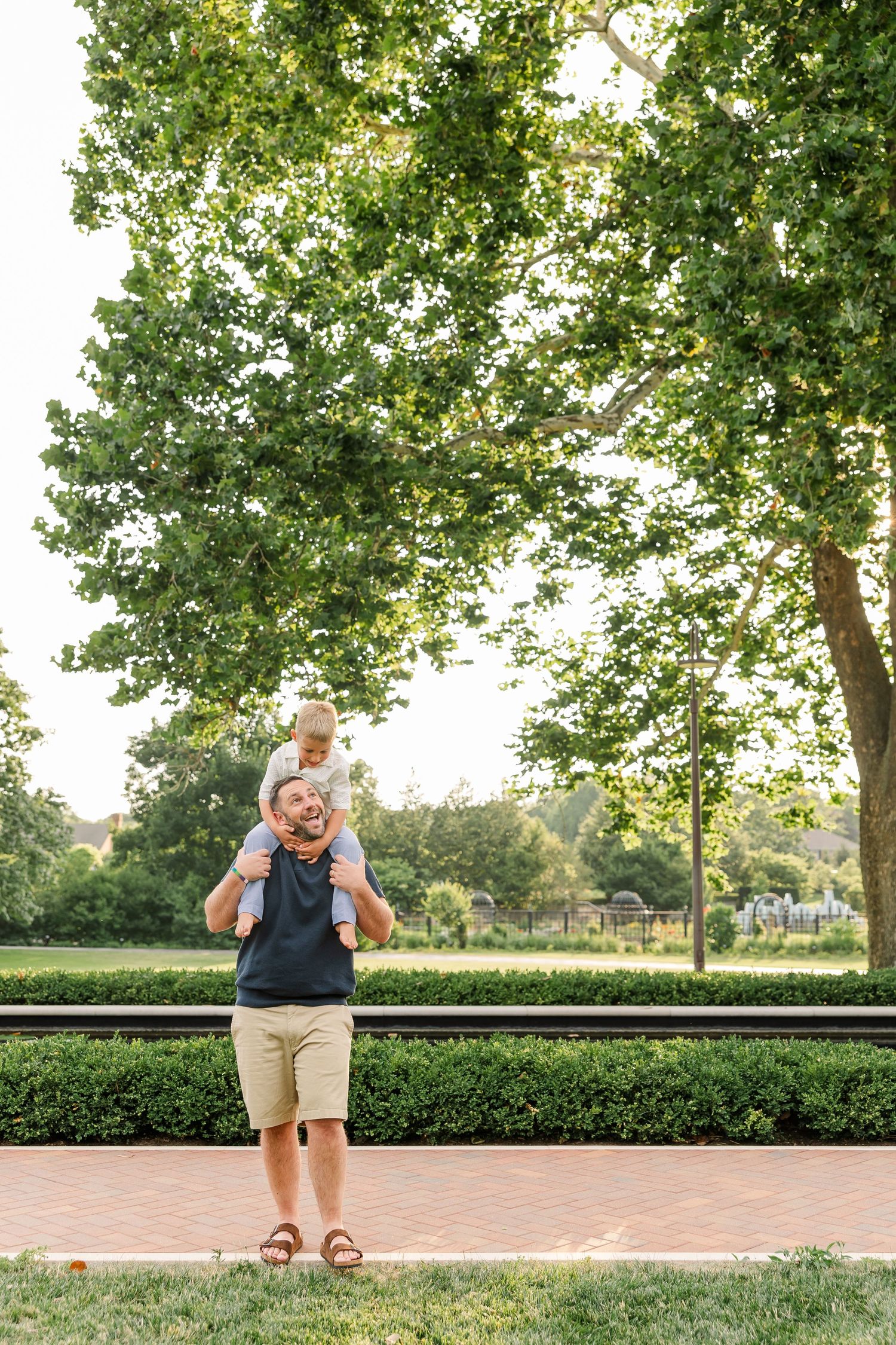 A father carries his young child on his shoulders in a lush green park on a sunny day.