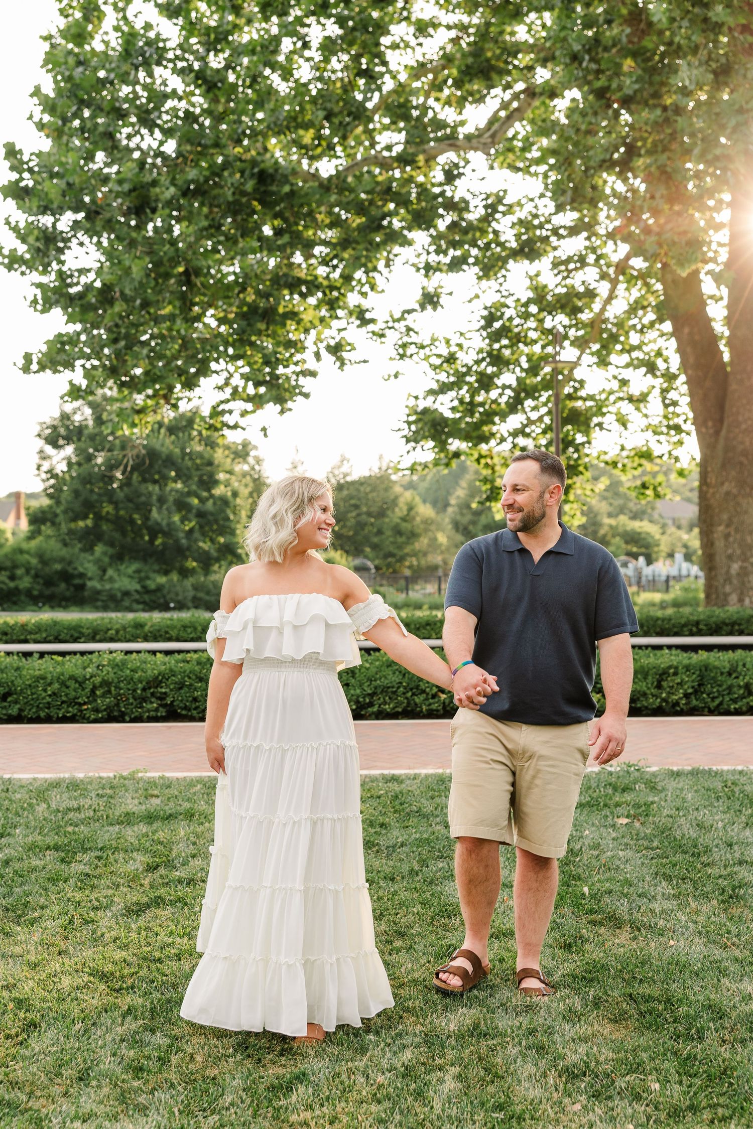 Couple holding hands and smiling at each other in sunny park, woman in white tiered dress, man in navy polo.