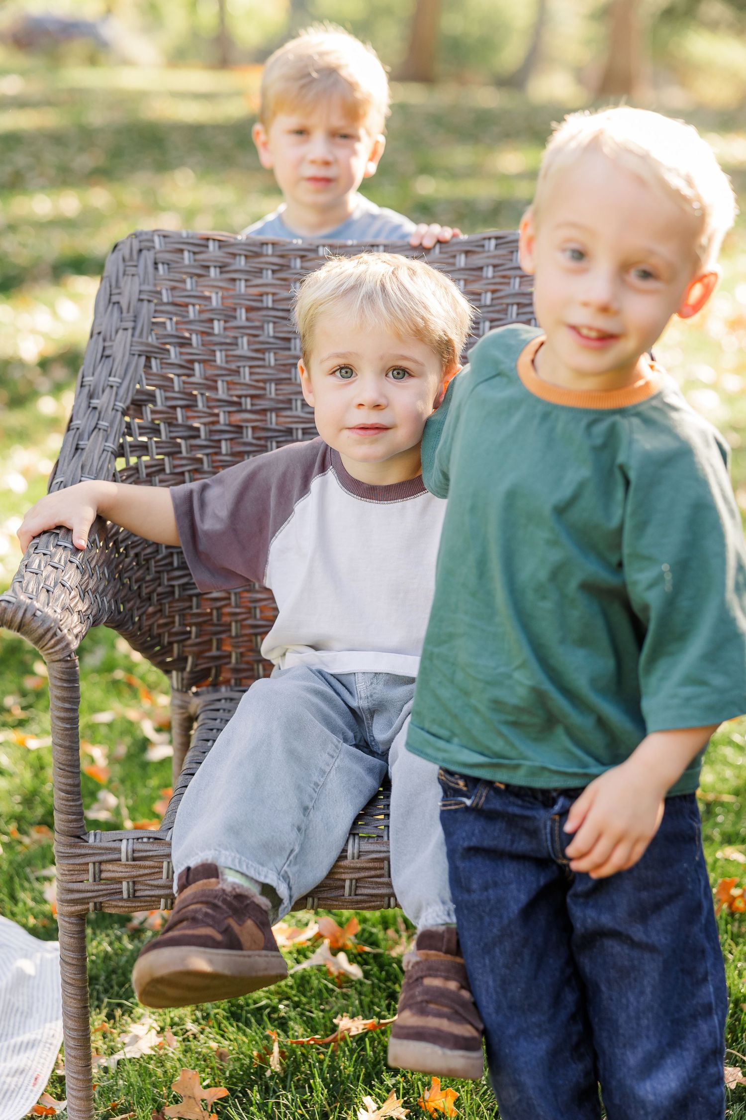 Three young blonde boys posing outdoors in a park, with one sitting on a wicker chair surrounded by fall leaves.