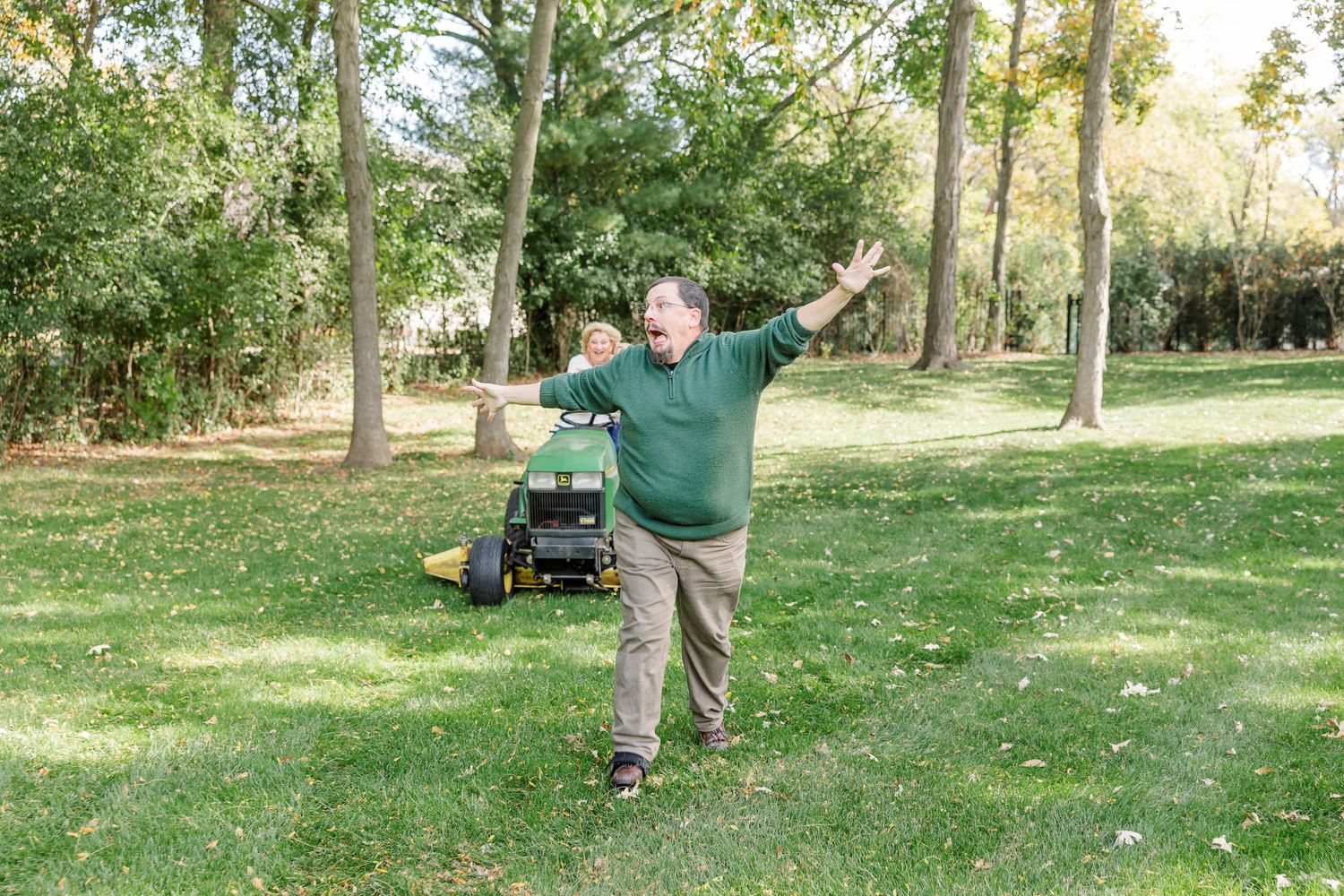 Cheerful man celebrating enthusiastically next to a green lawn mower in a tree-lined yard.