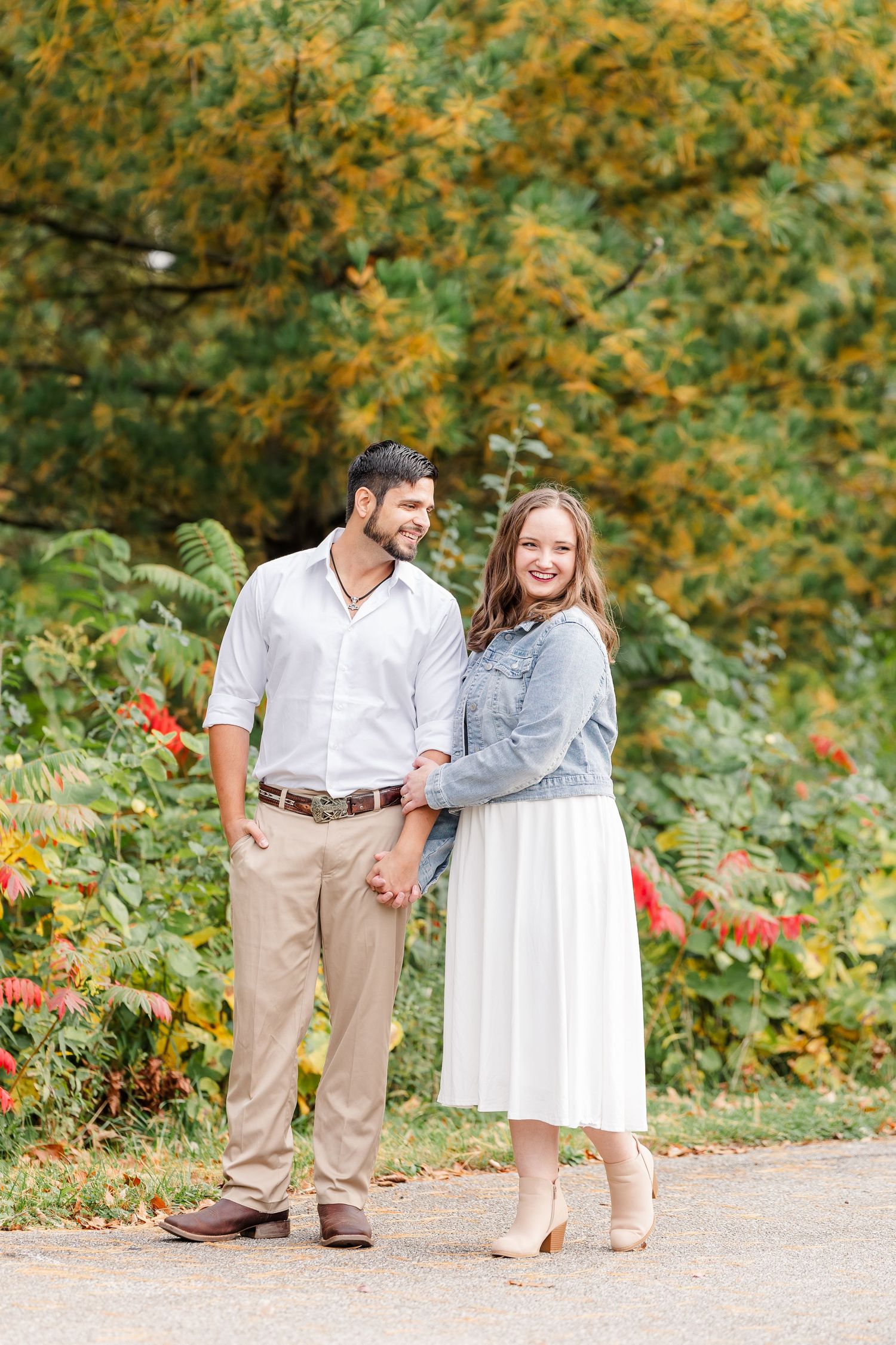 Smiling couple holding hands on a park path surrounded by colorful autumn foliage during an outdoor engagement photo session.