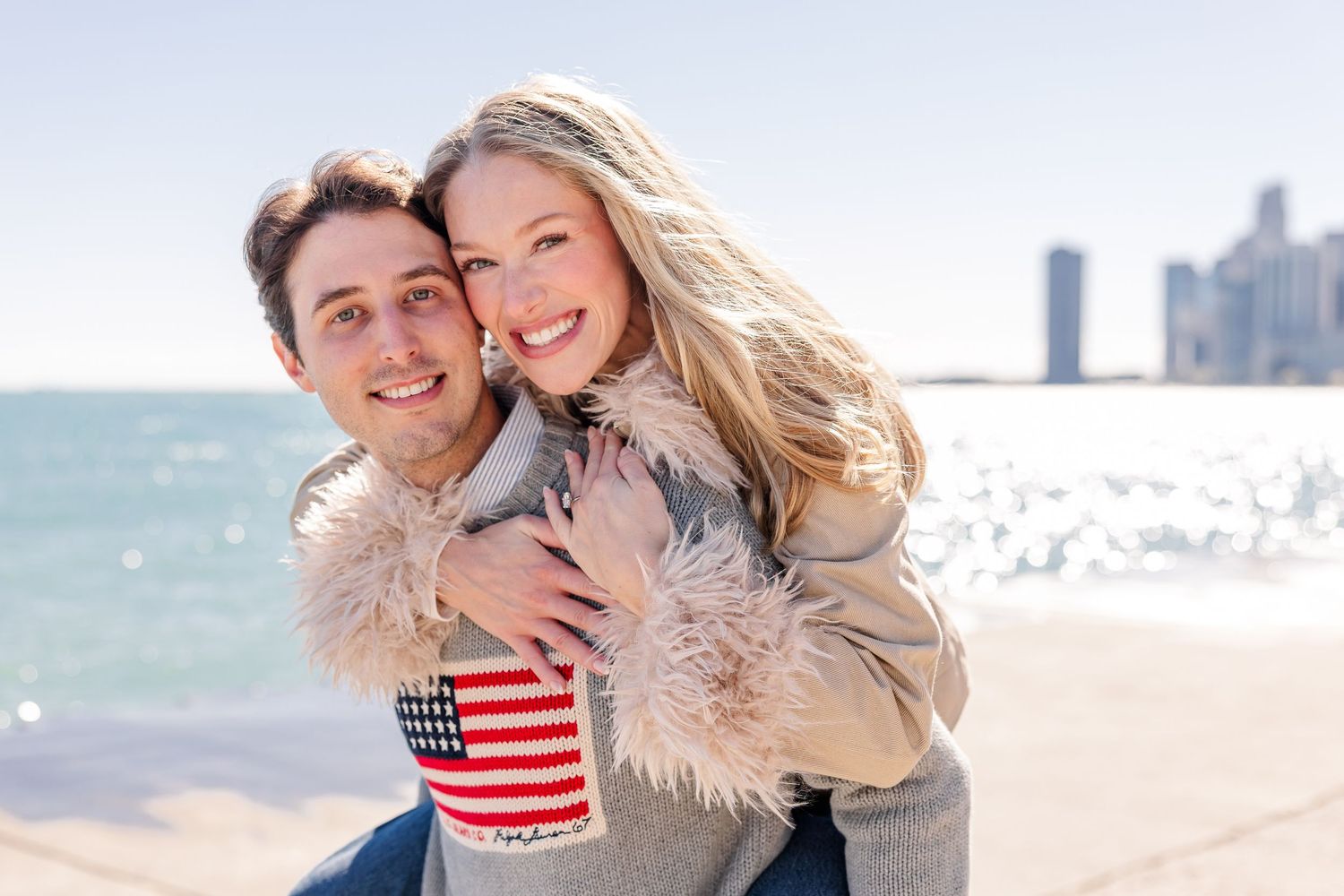 Happy couple sharing a piggyback ride on a sunny beach with city skyline, woman holding an American flag cup.