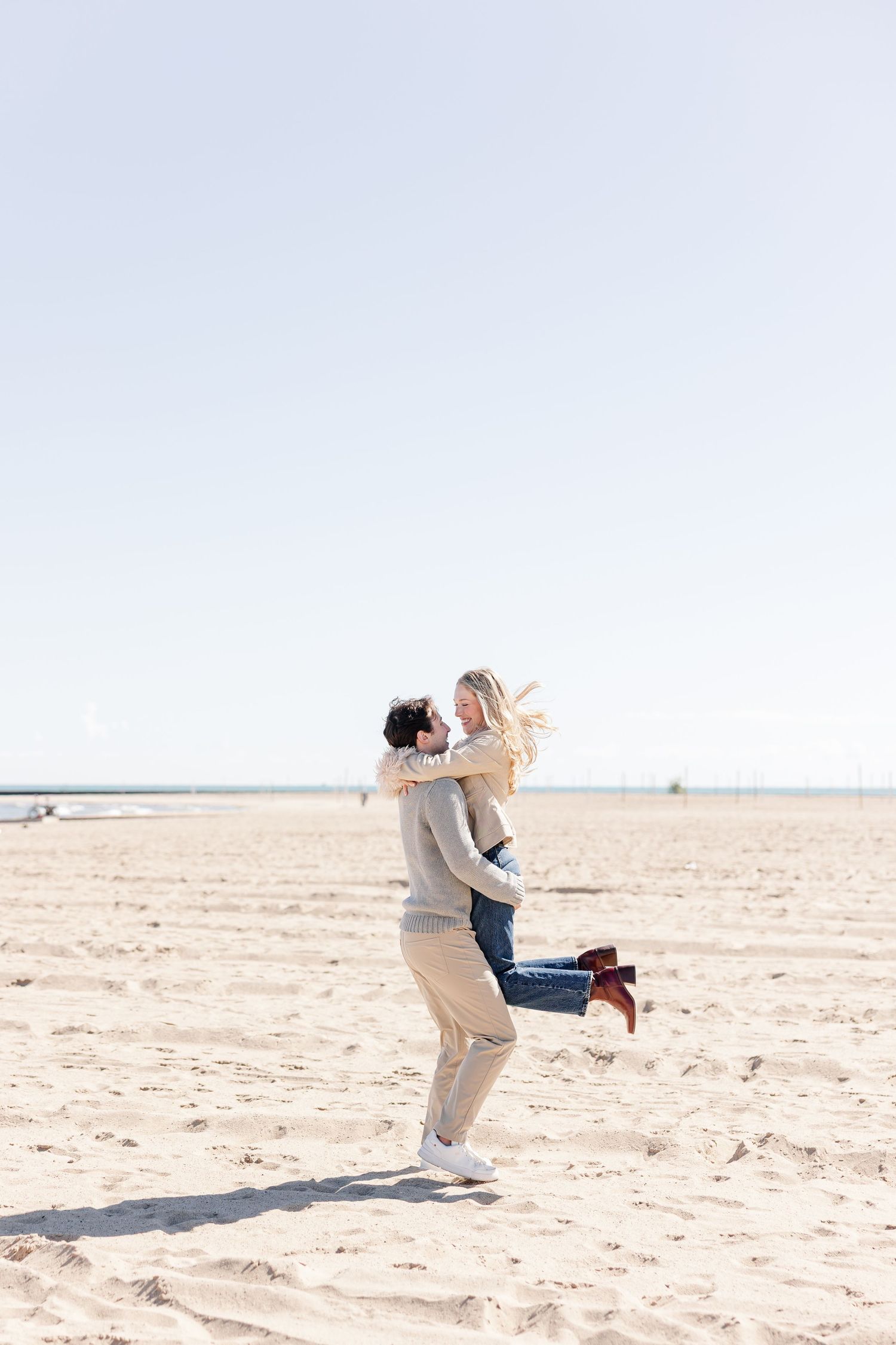 Joyful couple on a sandy beach, man lifting woman in the air while she laughs, both dressed in casual coastal outfits.