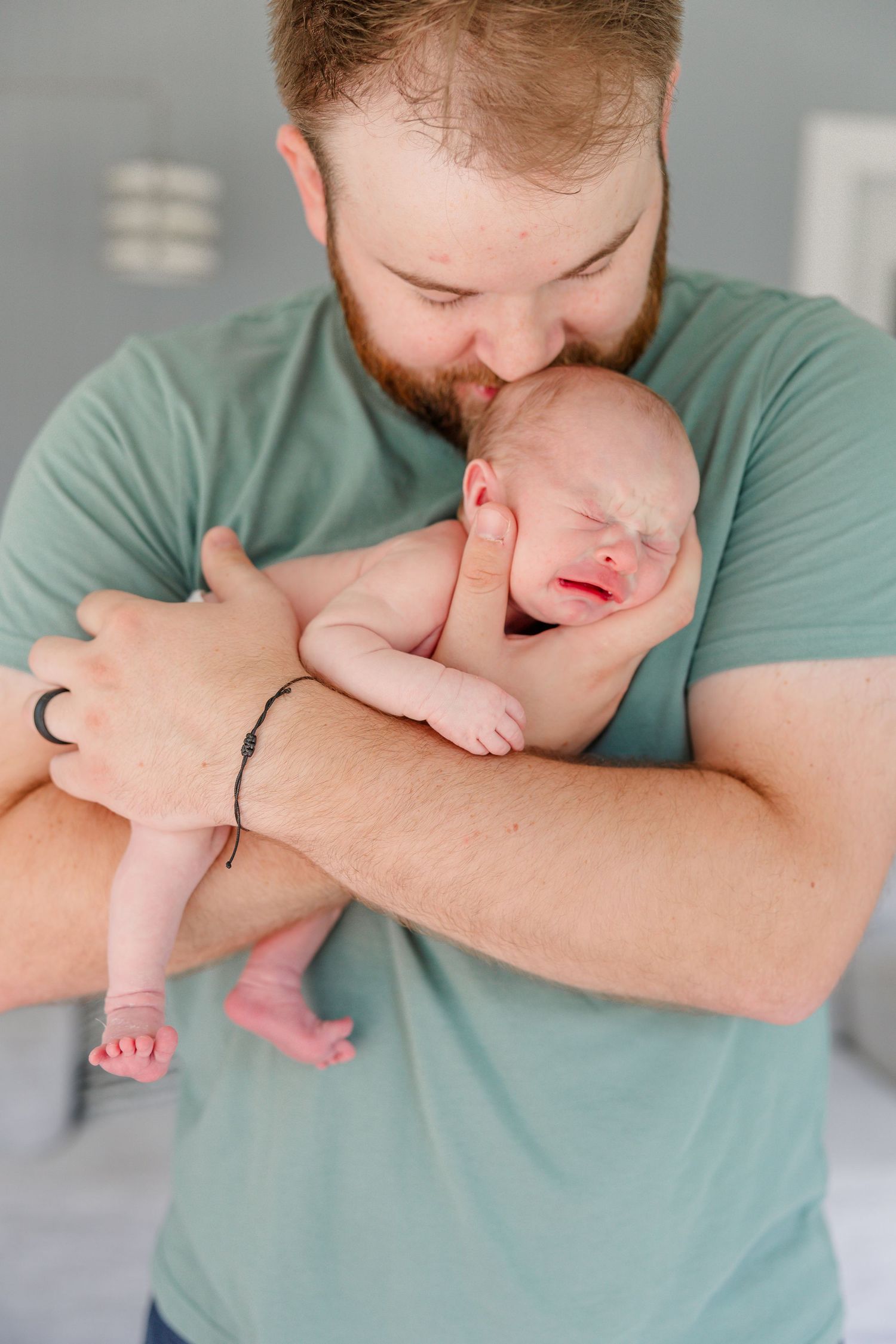 Loving bearded dad in teal shirt kisses his crying newborn baby while cradling them in his arms.
