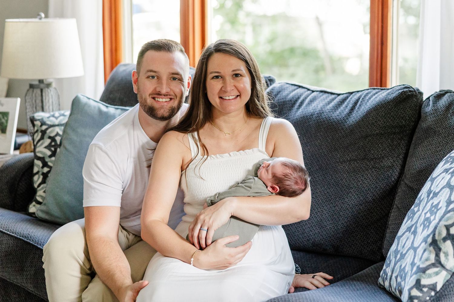 A smiling couple sits on a gray sofa holding their newborn baby in a bright, cozy living room.