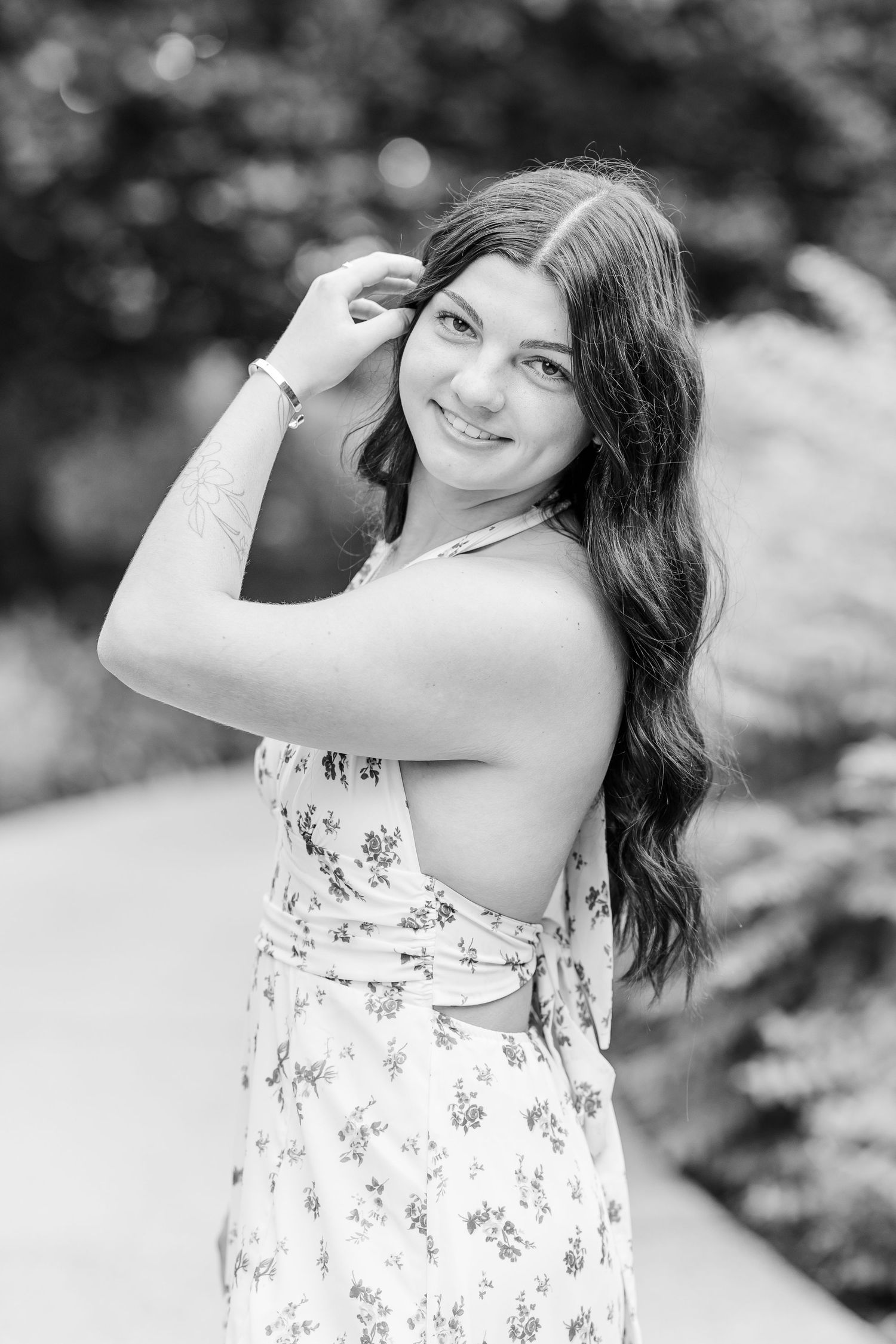 Black and white portrait of a smiling young woman in a floral dress posing outdoors among lush greenery.
